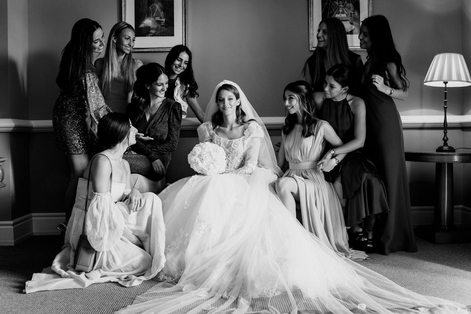 Black and white photo of a bride in a voluminous gown seated and surrounded by smiling bridesmaids in an elegant room.
