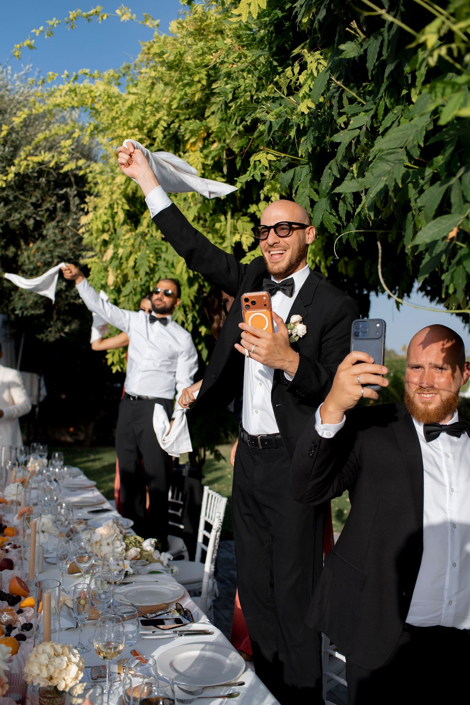 Groomsmen in black tuxedos wave white napkins and film with phones at a festive outdoor wedding reception table under leafy trees.
