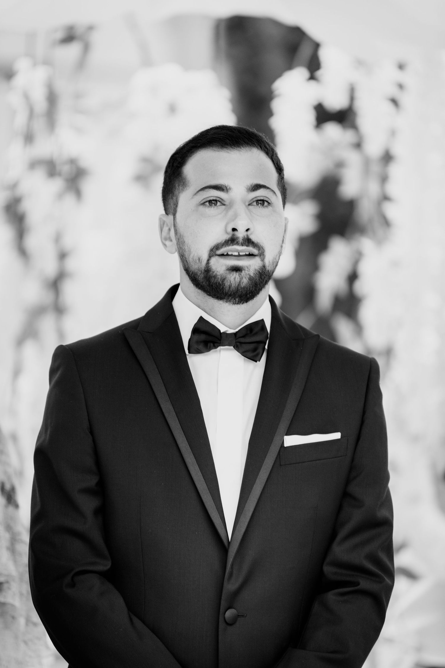Black and white portrait of a smiling groom with a beard wearing a tuxedo, bow tie and pocket square at a wedding.