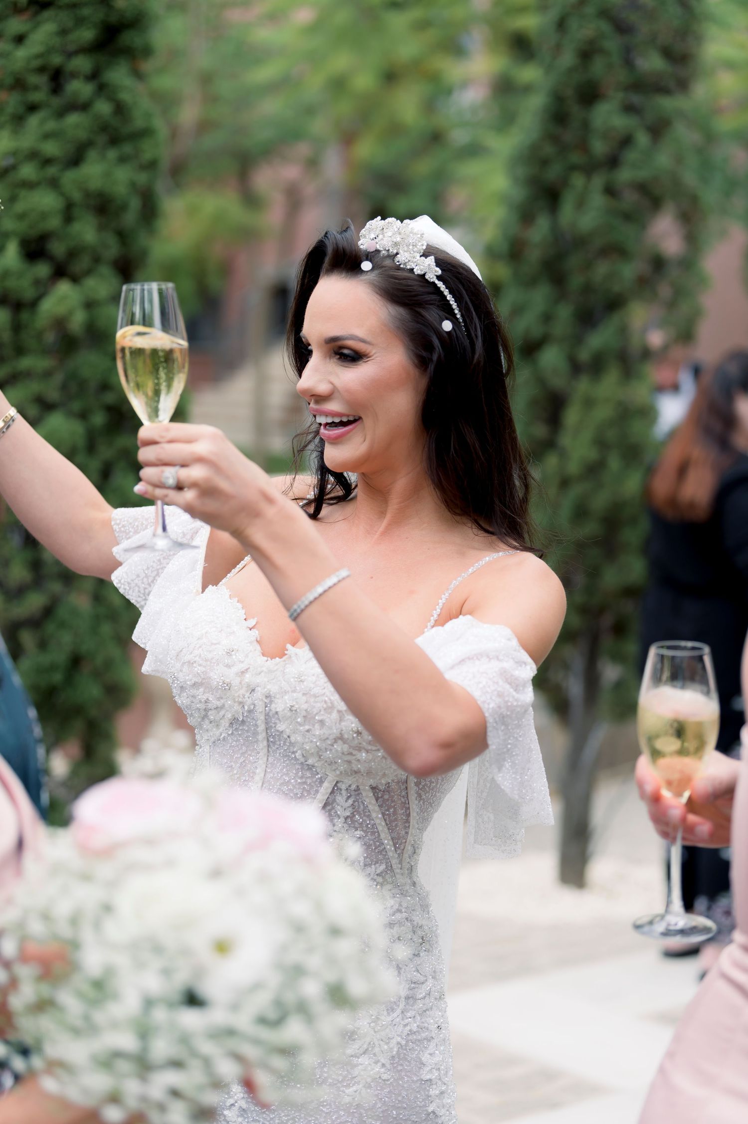 Smiling bride in a white lace gown and floral tiara raises a champagne glass in a toast outdoors at her wedding celebration.