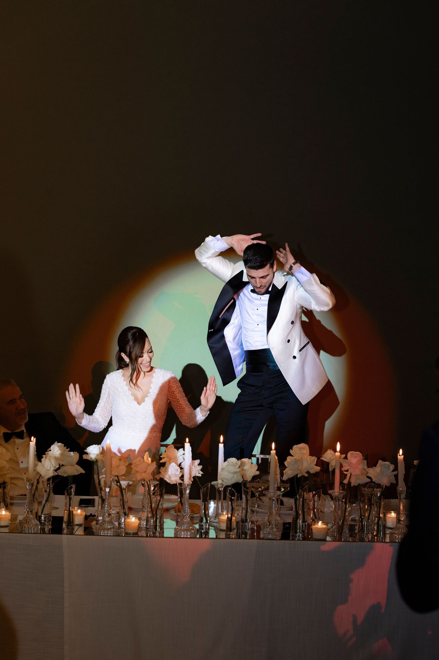 A couple in formal wedding attire dances on a candlelit reception table under a warm spotlight, surrounded by flowers and glowing candles.