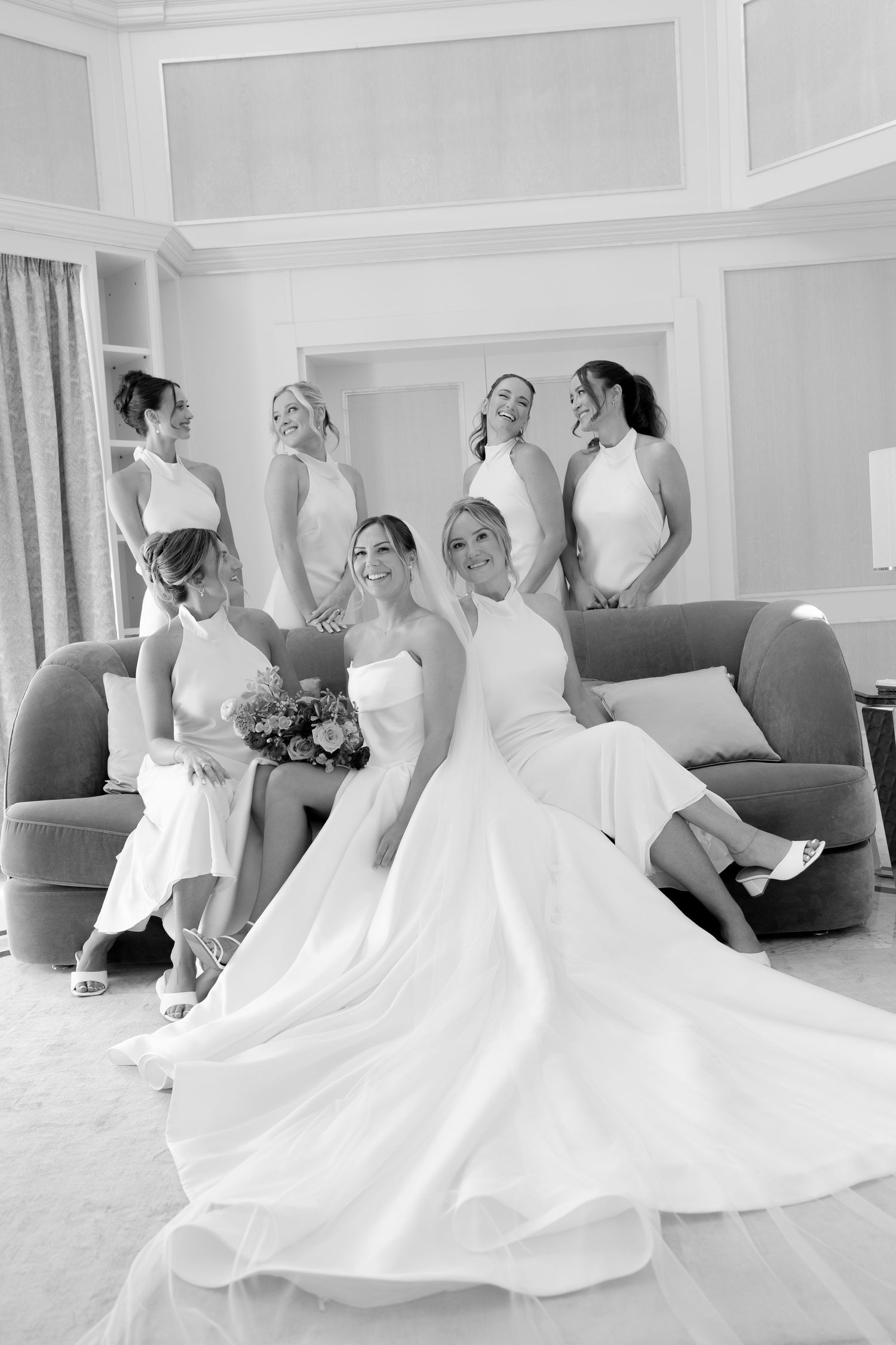Black and white photo of a bride in a flowing gown seated on a sofa with six bridesmaids in white dresses posing around her.