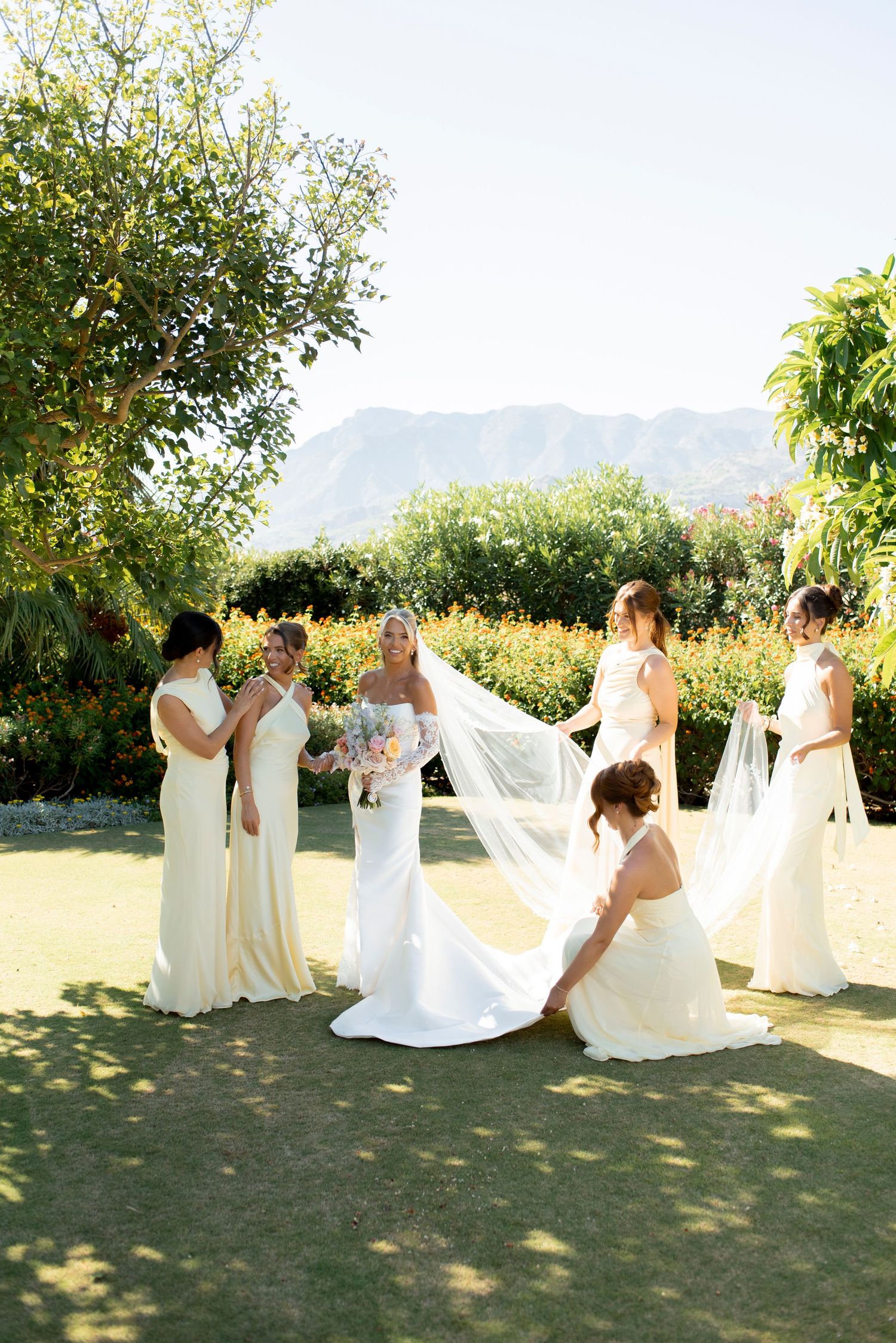 Bride in a flowing white gown with a long veil holding a bouquet, surrounded by four bridesmaids in cream dresses on a sunny garden lawn.