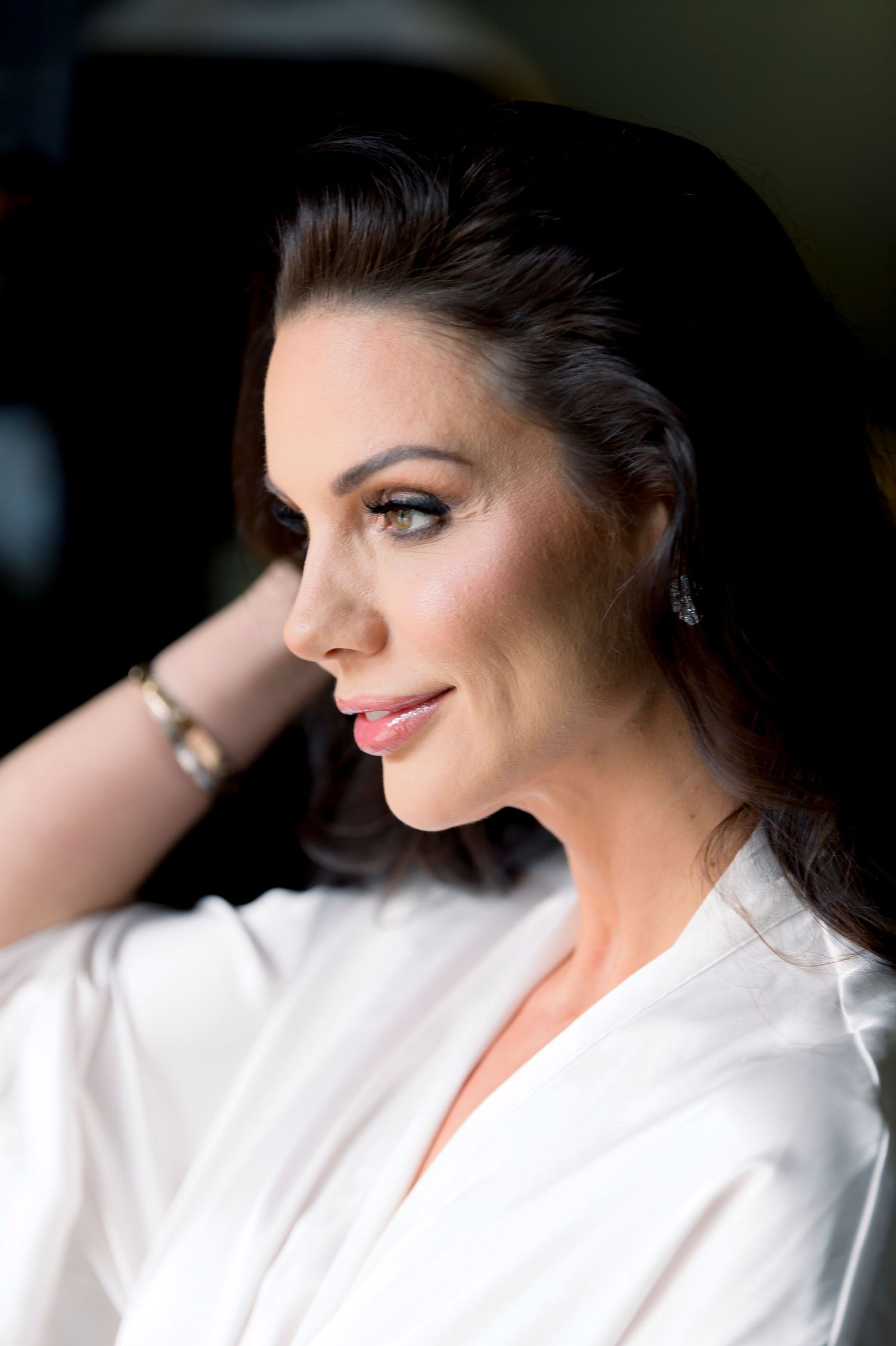 Side profile of a woman with dark hair swept back, wearing a white satin robe, pink lipstick and a gold bracelet near a window.