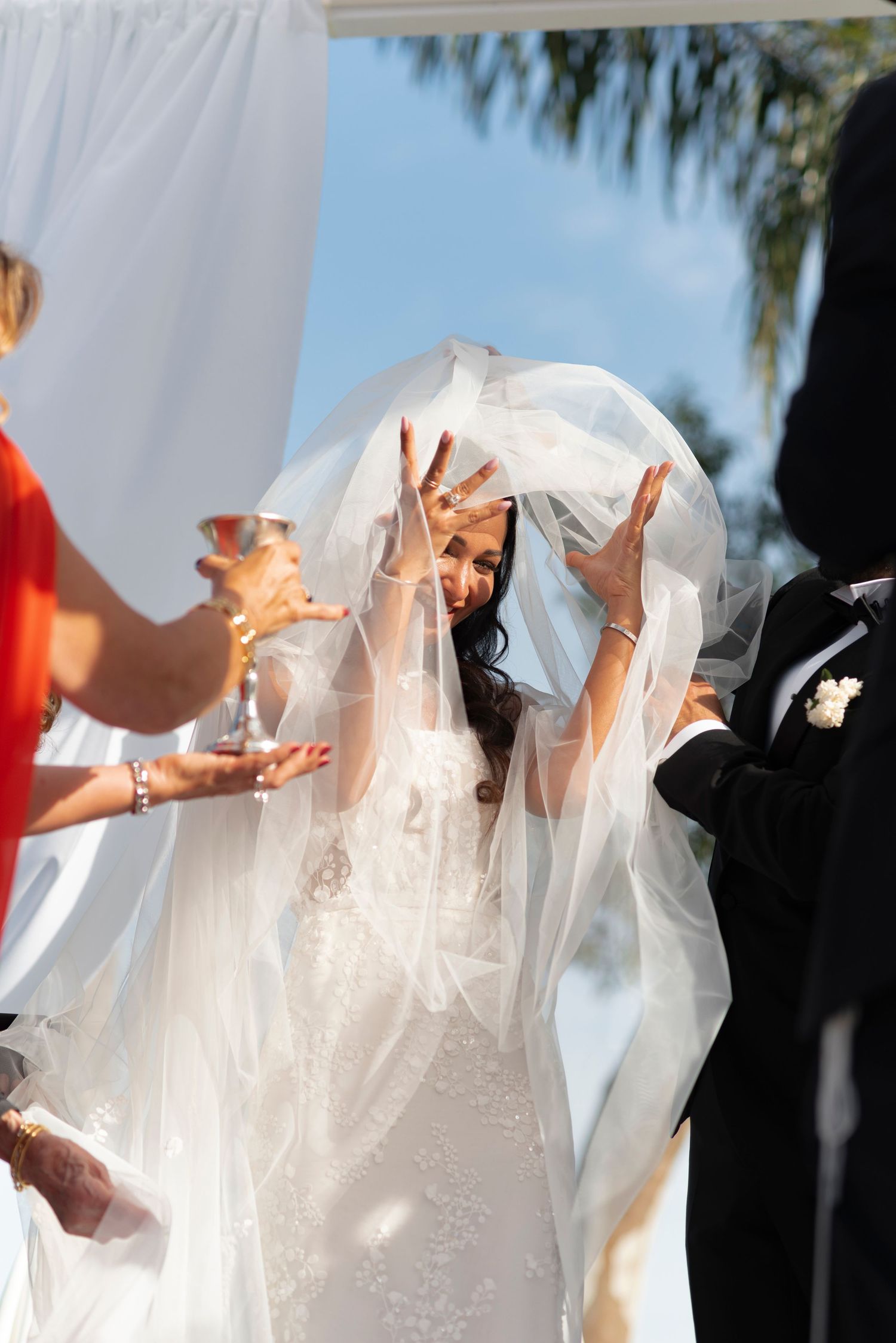 Bride in a lace wedding gown and sheer veil raises her hands joyfully under a canopy during an outdoor ceremony as a guest holds a goblet.