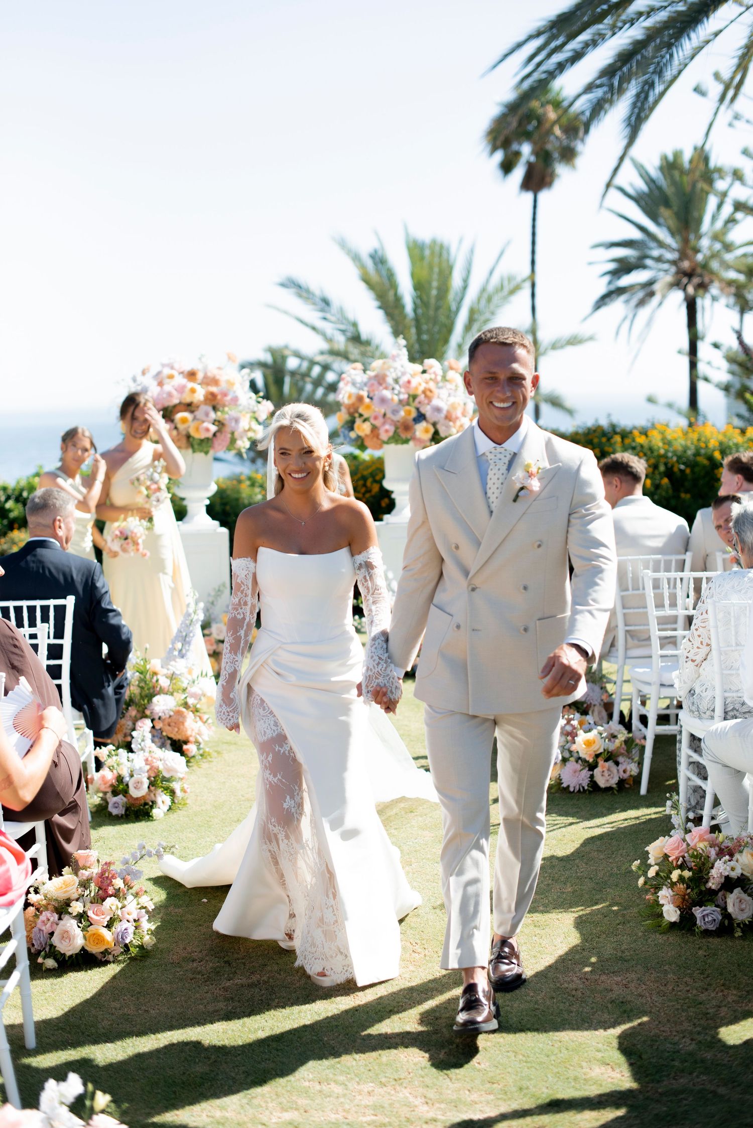 Smiling bride in a strapless white lace gown walks hand in hand with a groom in a beige suit through an outdoor garden ceremony.