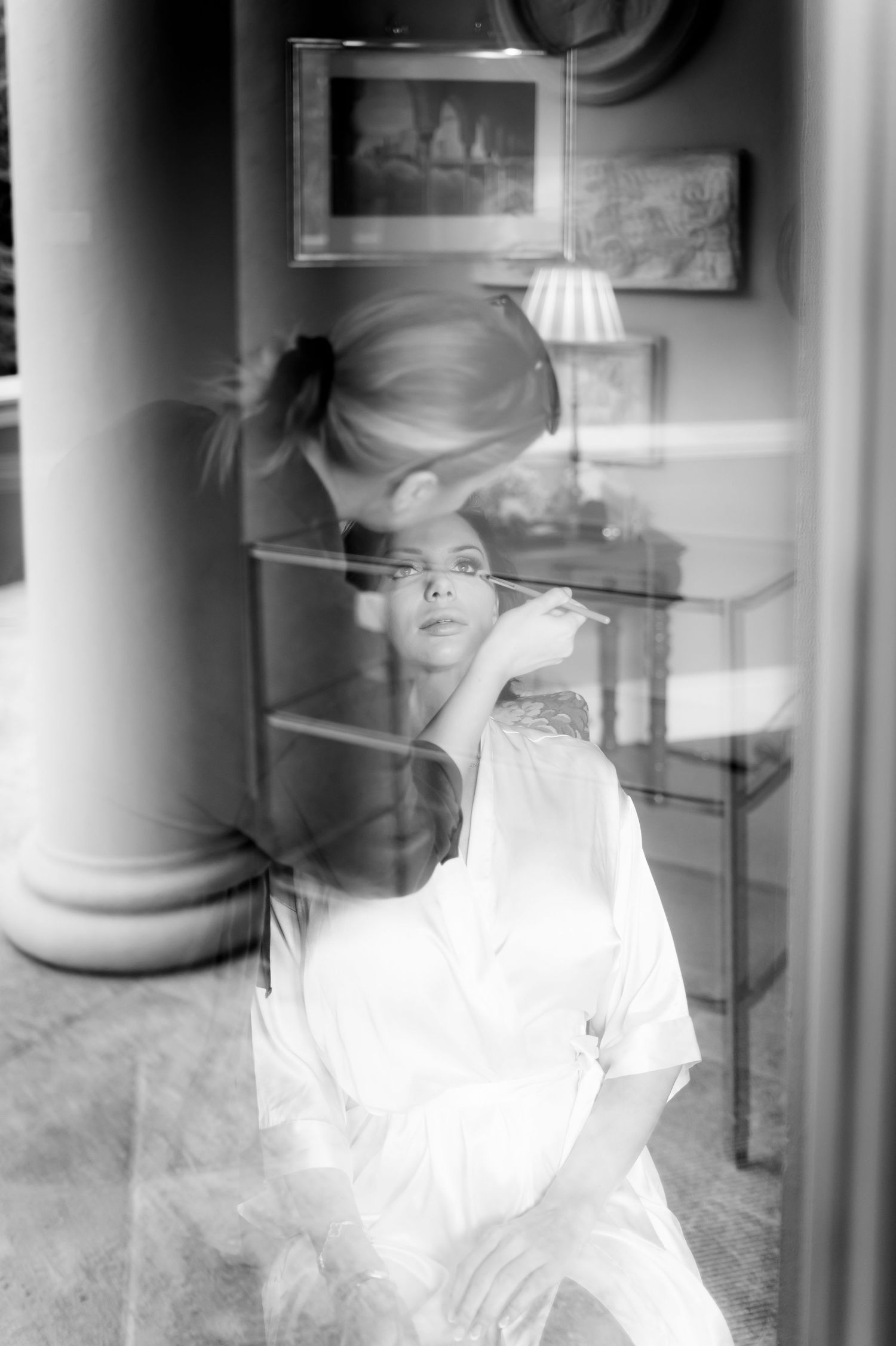 Black and white photo seen through a window of a makeup artist applying makeup to a woman in a satin robe in an elegant room.