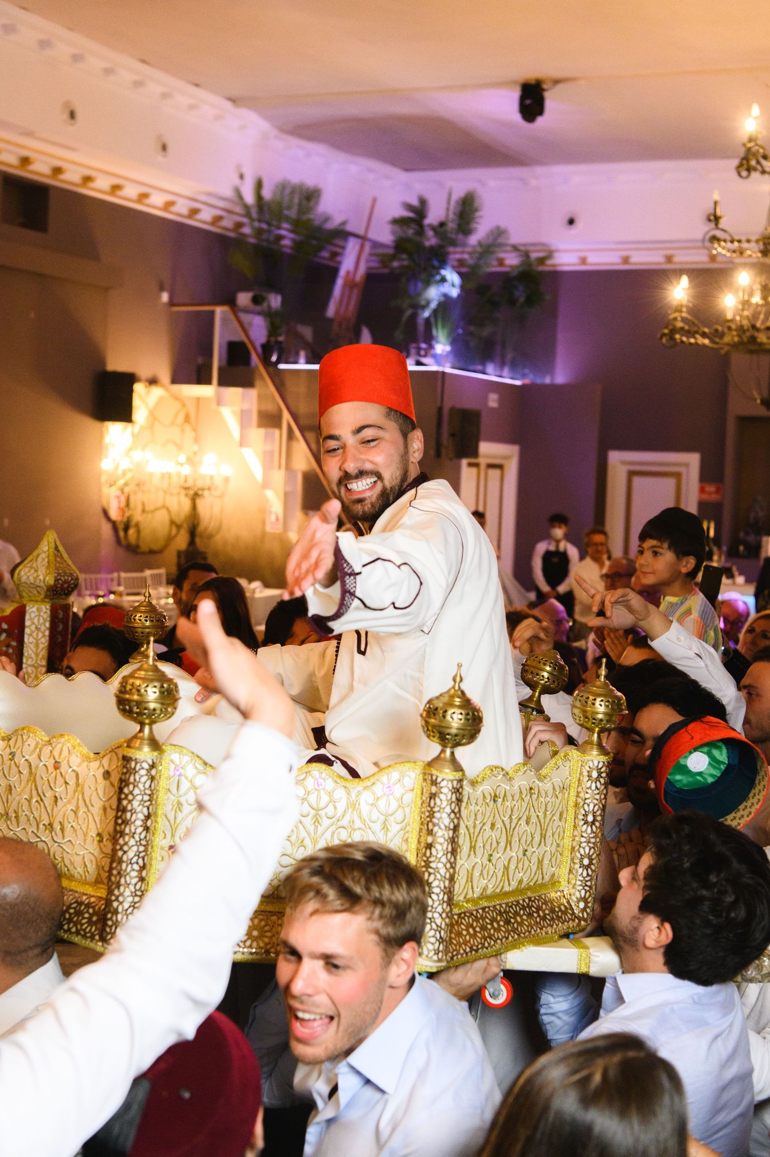 Groom in a red fez and white outfit is lifted on a golden ornate chair by cheering guests at a lively wedding celebration.
