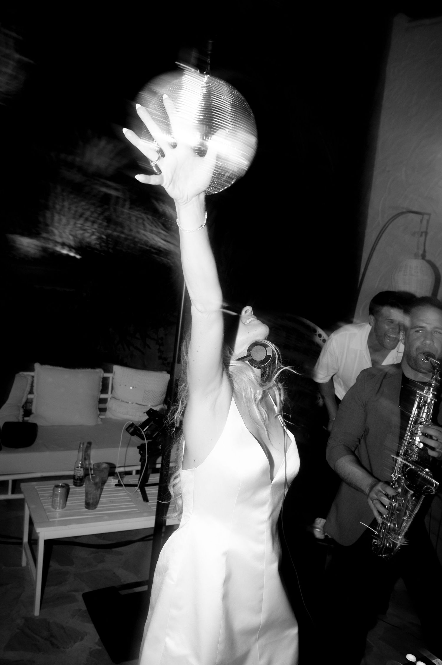 Woman in a white dress reaches toward a mirrored disco ball at a party while a saxophonist plays in the background, in black and white.