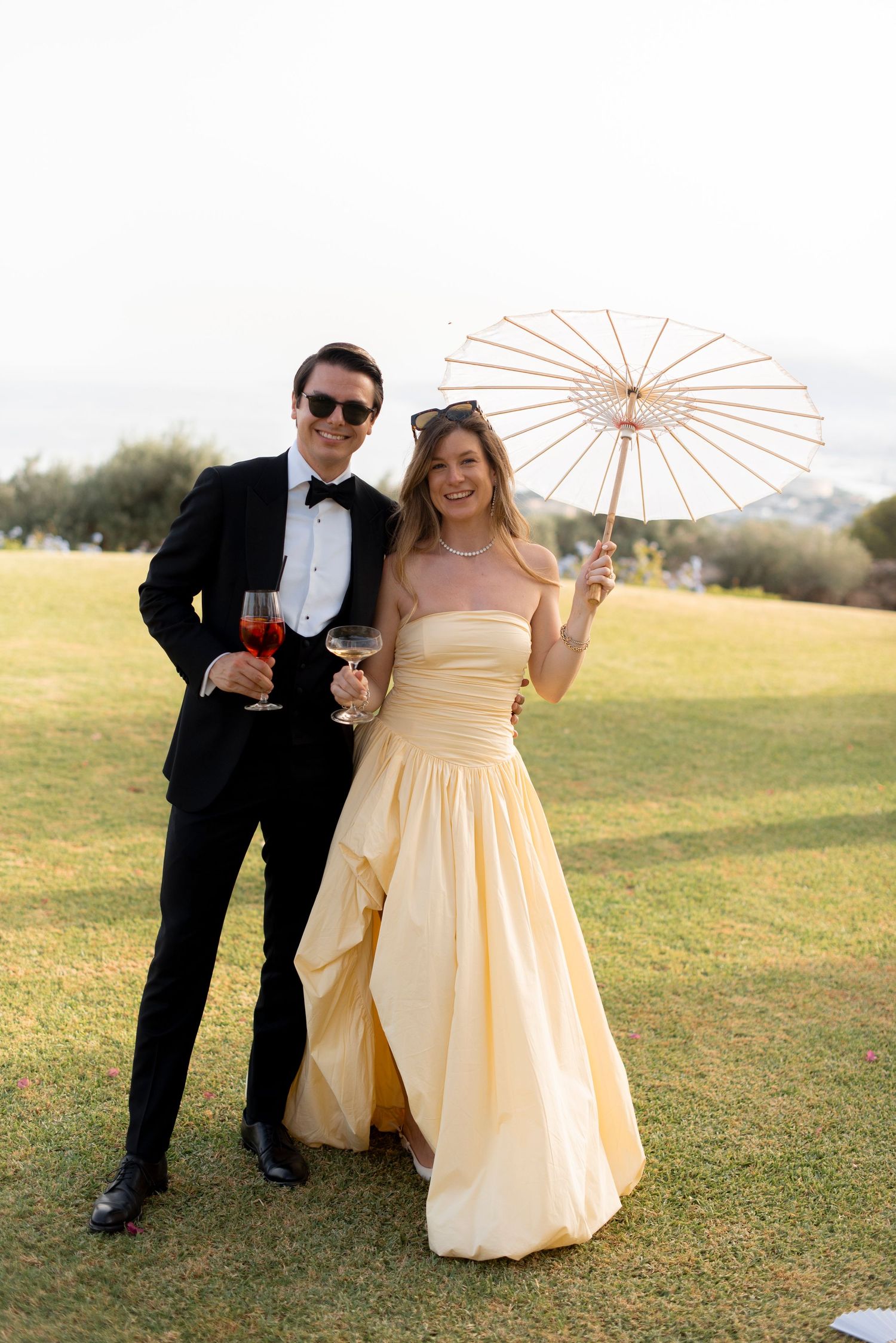 Smiling couple at an outdoor wedding, he in a black tuxedo and she in a yellow gown holding a white parasol, both with drinks on a lawn.