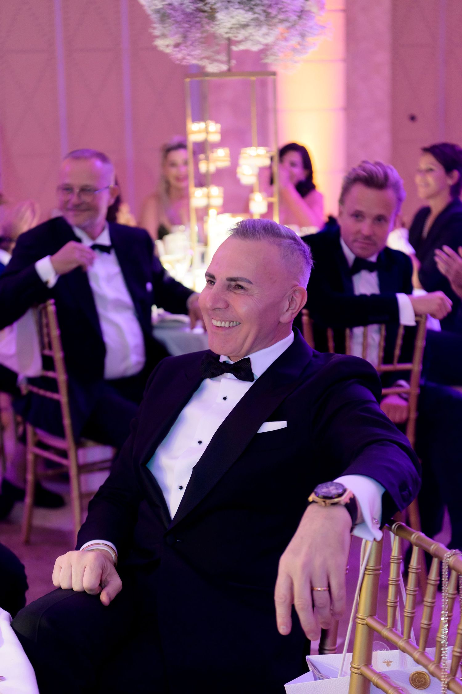 Smiling man in a black tuxedo and bow tie sits at an elegant wedding reception, with guests and glowing pink lighting behind him.