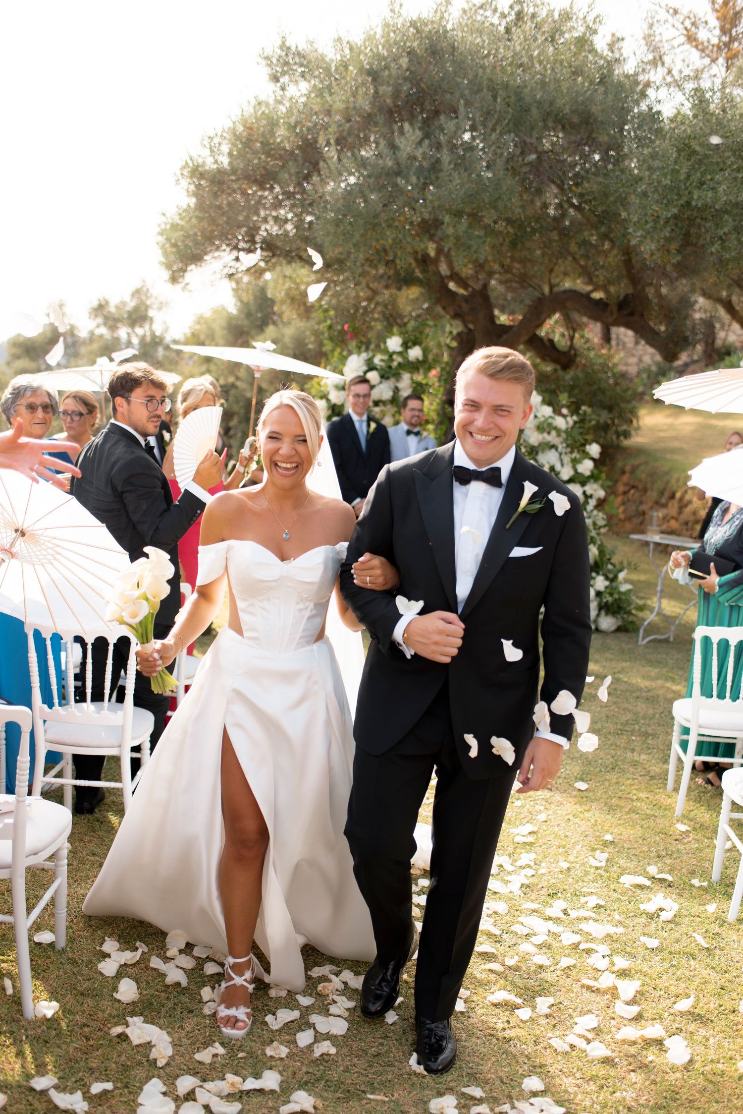 Smiling bride and groom in a black tuxedo walk down the aisle through falling white petals as wedding guests cheer in an outdoor garden.
