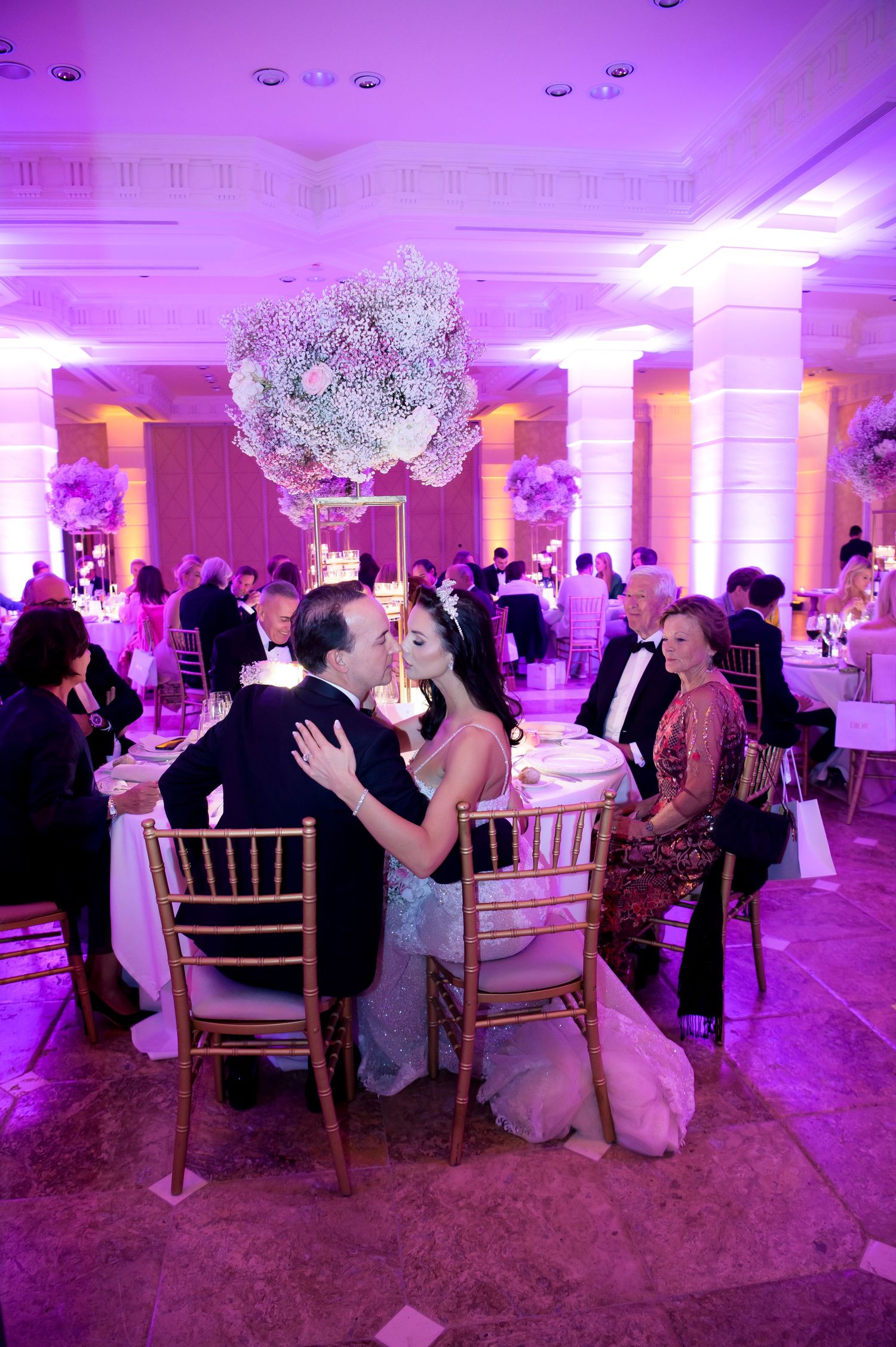 A couple shares a kiss at an elegant wedding reception with purple lighting, floral centerpieces, and guests seated at tables.