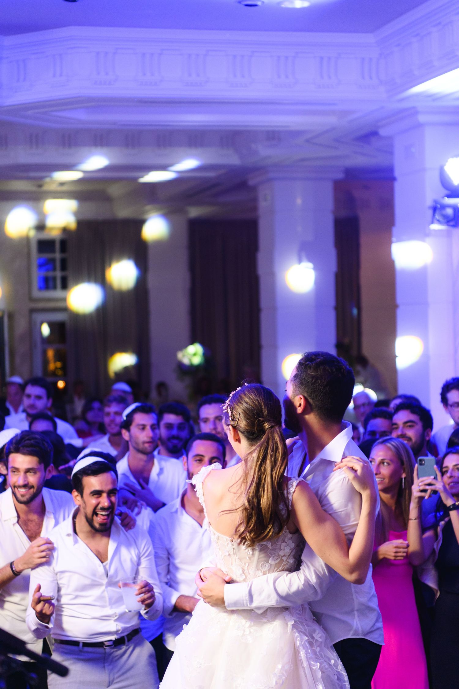 Bride and groom share a first dance in a grand ballroom while cheering wedding guests watch and clap around them.