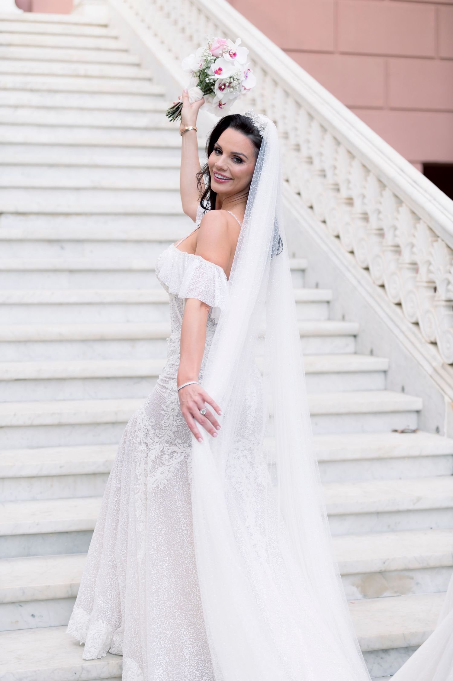 Smiling bride in an off-shoulder lace gown and long veil holds a pink bouquet while posing on a grand white staircase.