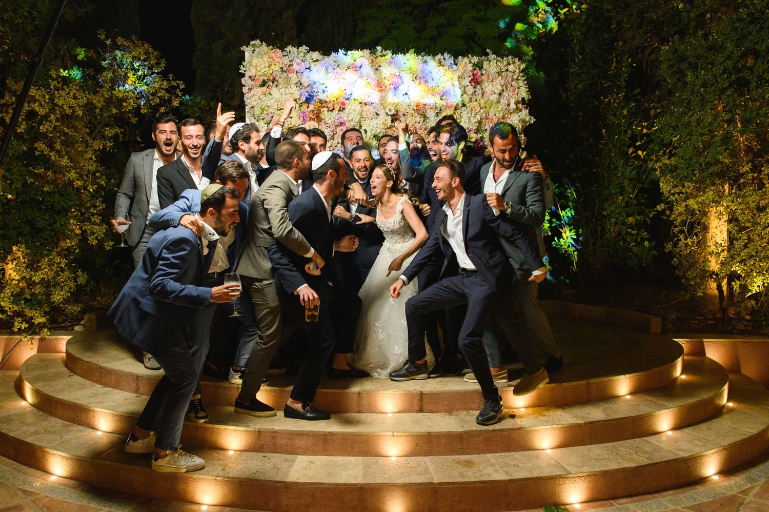 Wedding guests in formal attire celebrate and cheer around a bride and groom on a tiered circular stage with a floral backdrop at night.