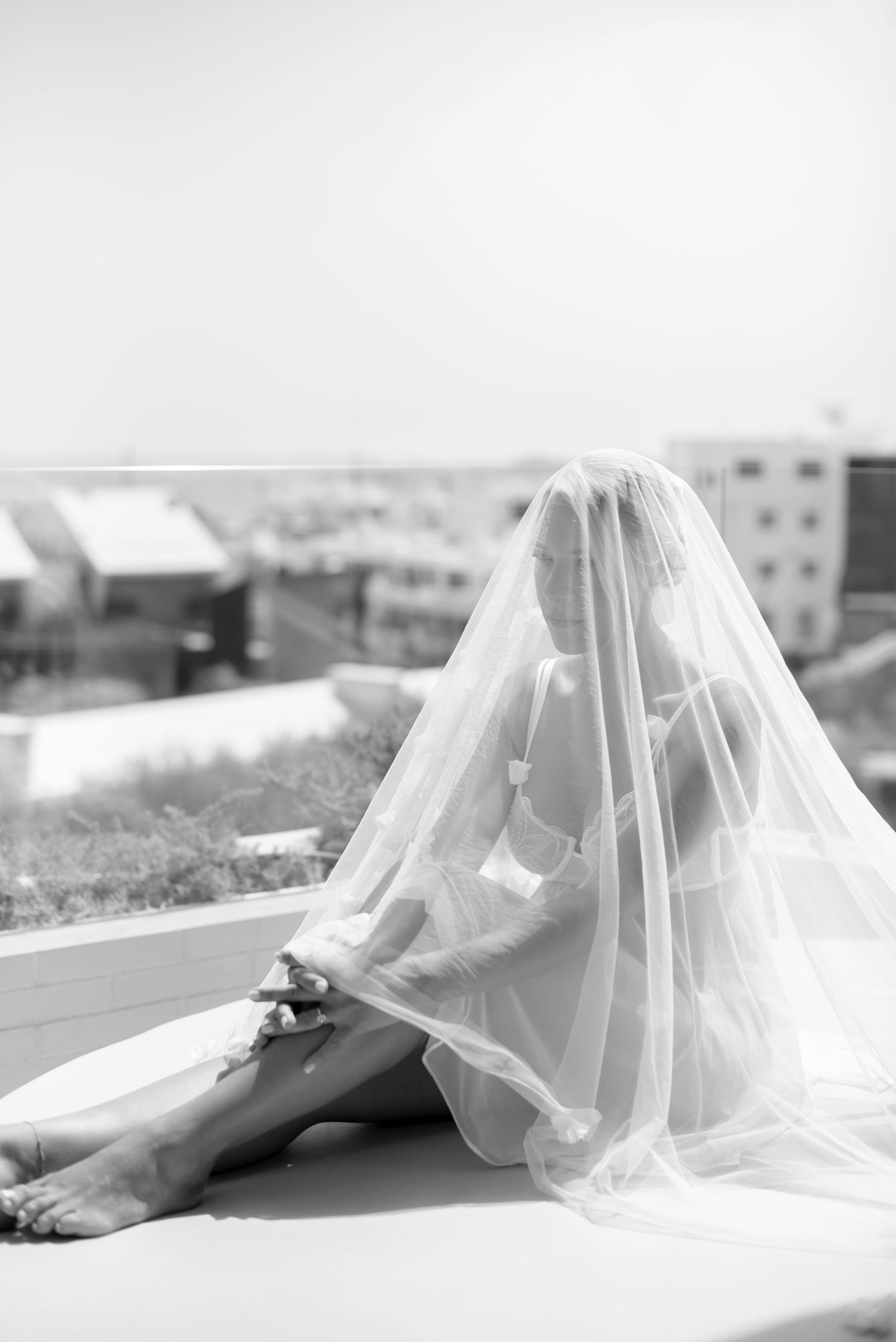 Black and white photo of a bride seated on a rooftop ledge, covered by a sheer tulle veil, with a city skyline behind her.