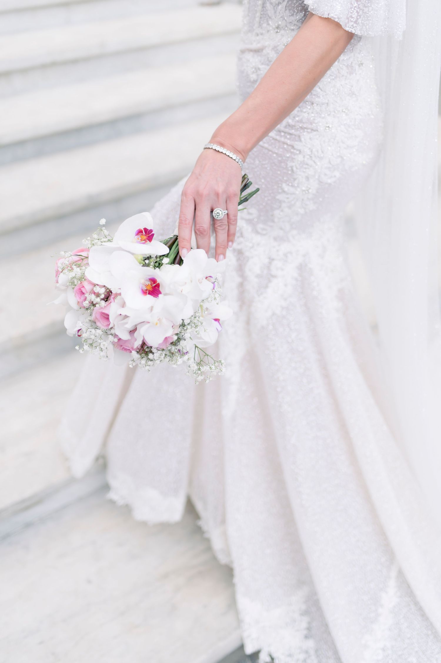 Bride in a lace wedding gown holds a bouquet of white and pink orchids with baby's breath on stone steps.