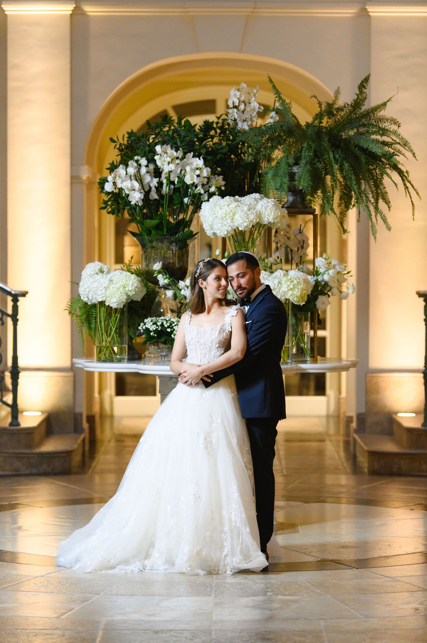 Bride in a flowing tulle gown and groom in a navy suit embrace in an elegant arched hall with white floral arrangements.