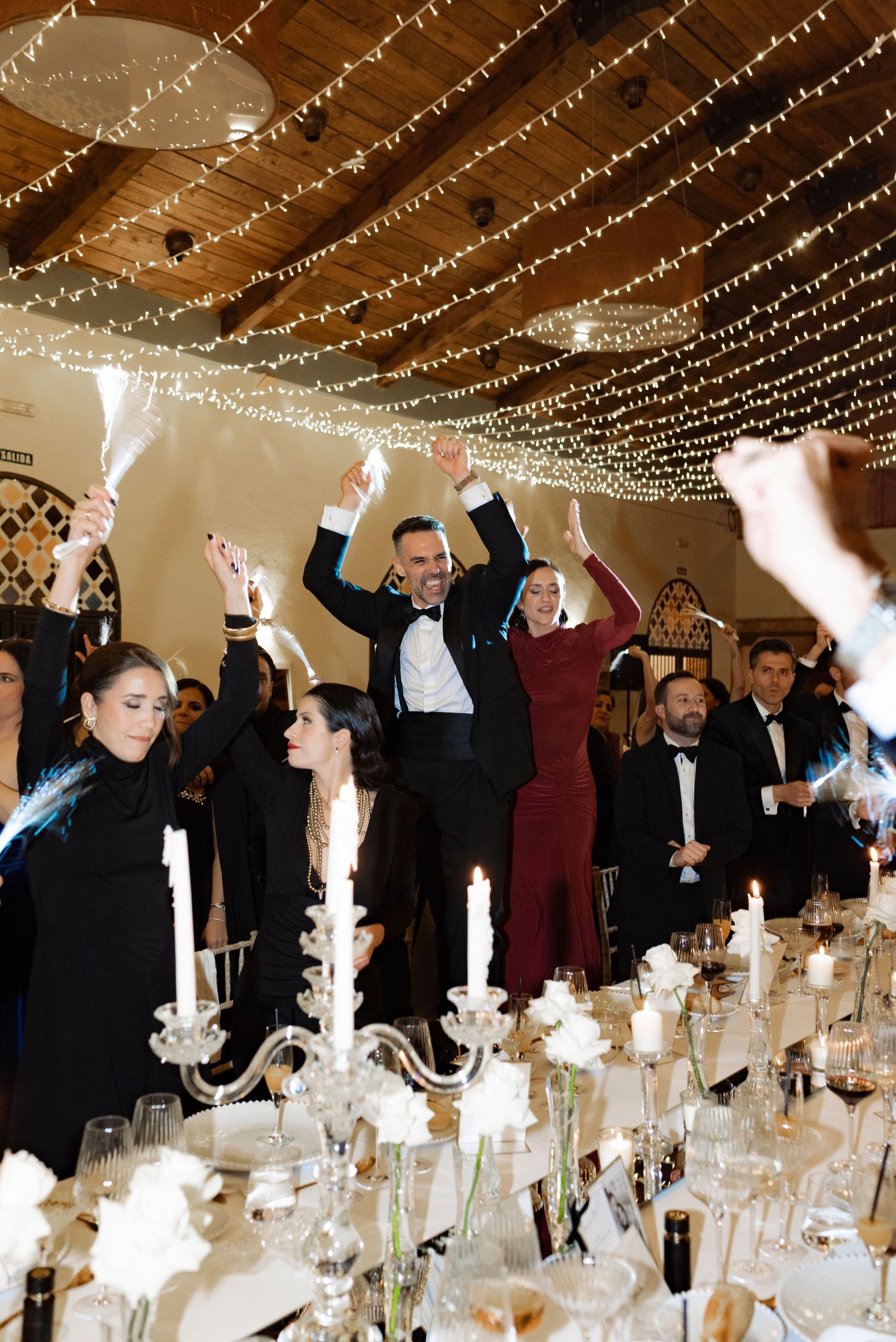 Wedding guests in formal attire cheer and raise sparklers at a candlelit reception table beneath a ceiling of twinkling string lights.