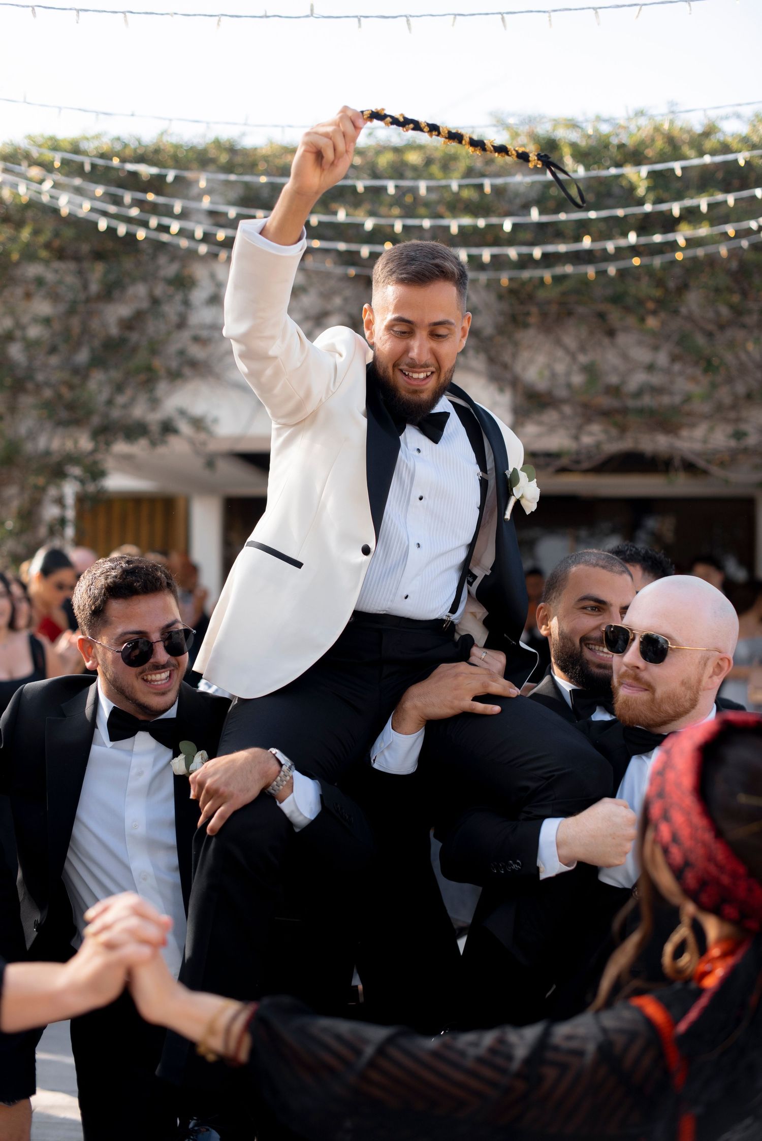 Smiling groom in a white tuxedo jacket and bow tie is lifted on friends' shoulders while waving a scarf at an outdoor wedding celebration.