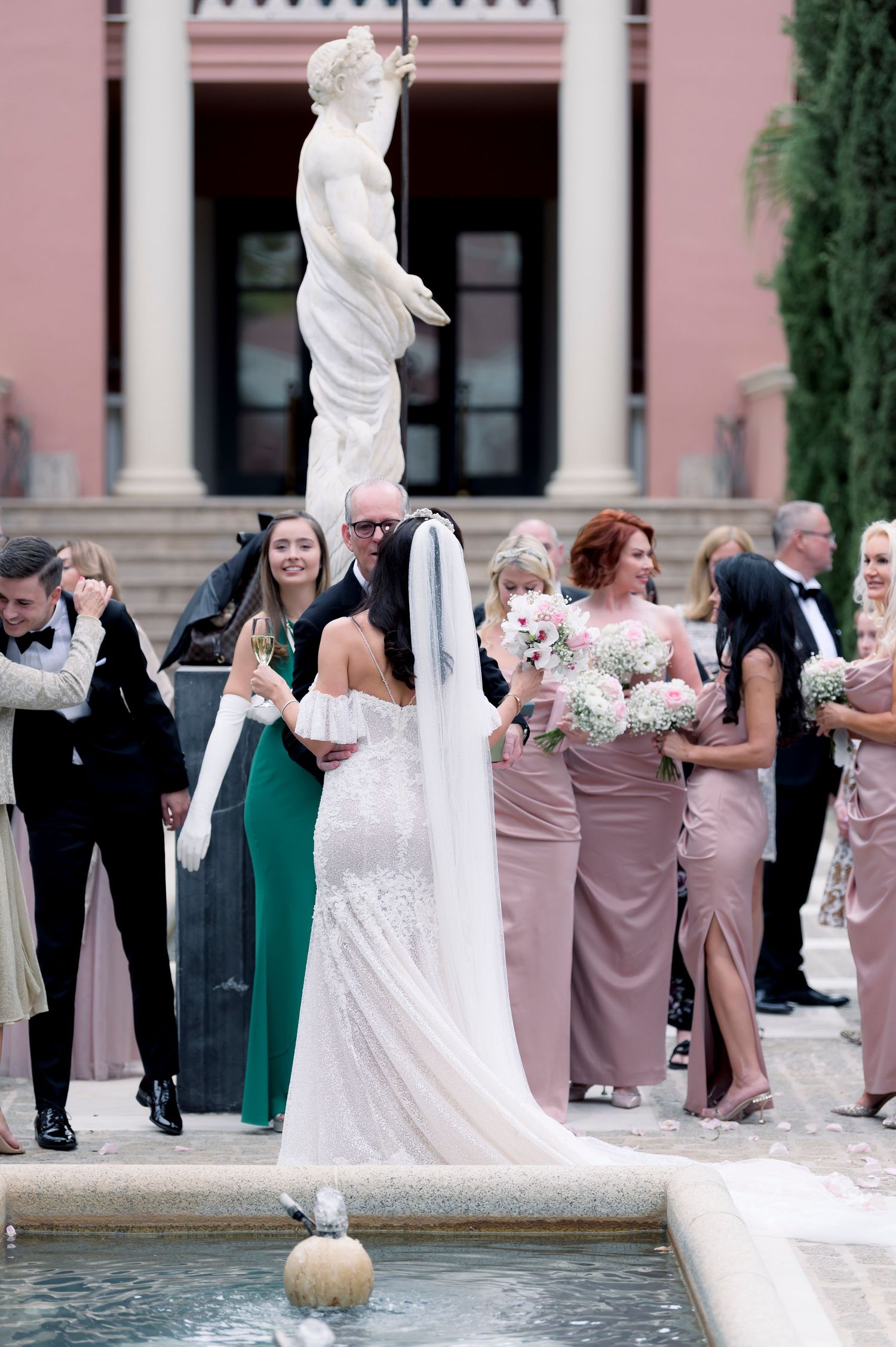 Bride in a lace gown with long veil greets bridesmaids in blush dresses holding bouquets beside a fountain and statue at a wedding.