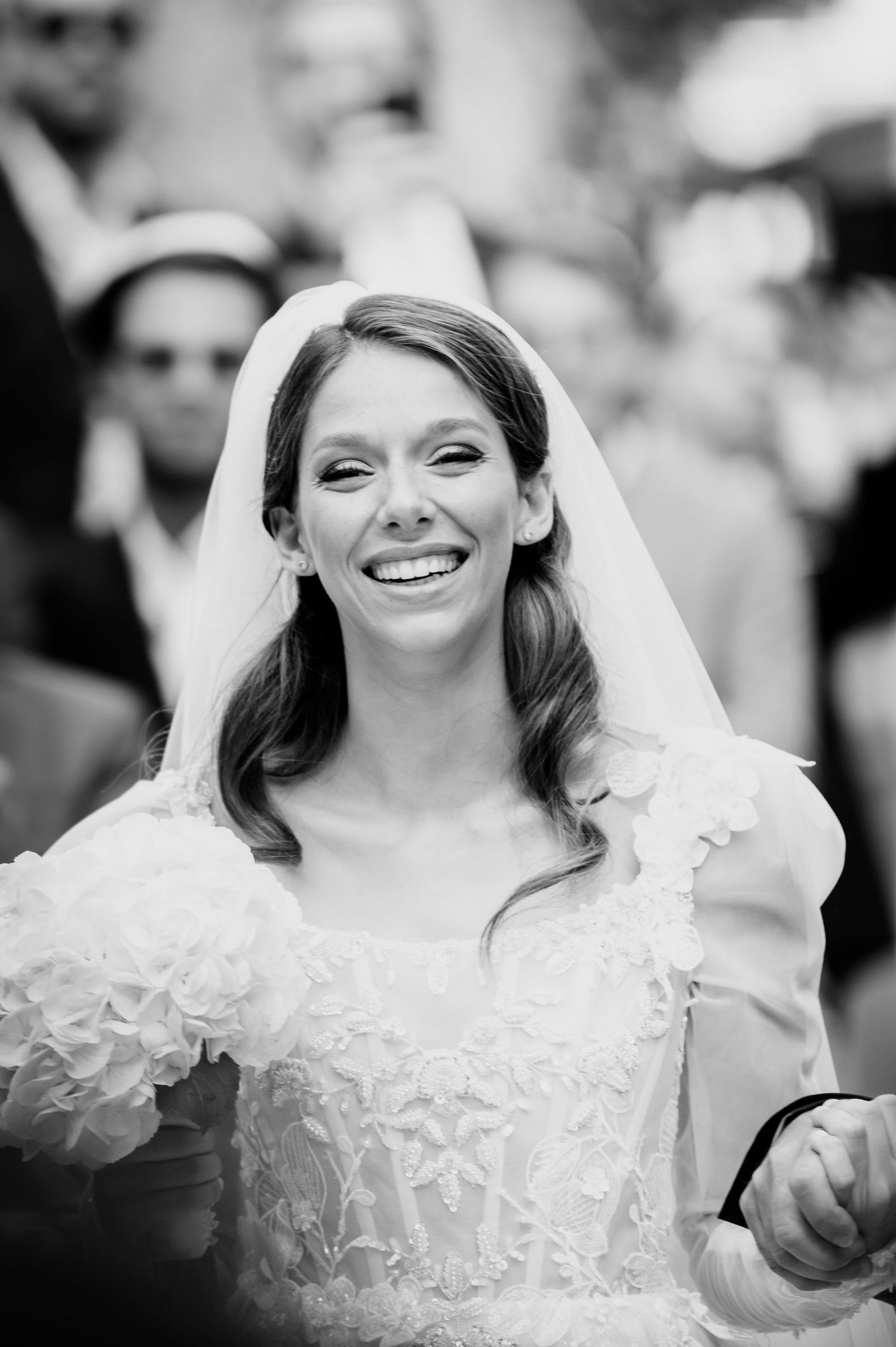 Smiling bride in a lace wedding gown with puffed sleeves and a sheer veil walks hand in hand through a crowd in black and white.