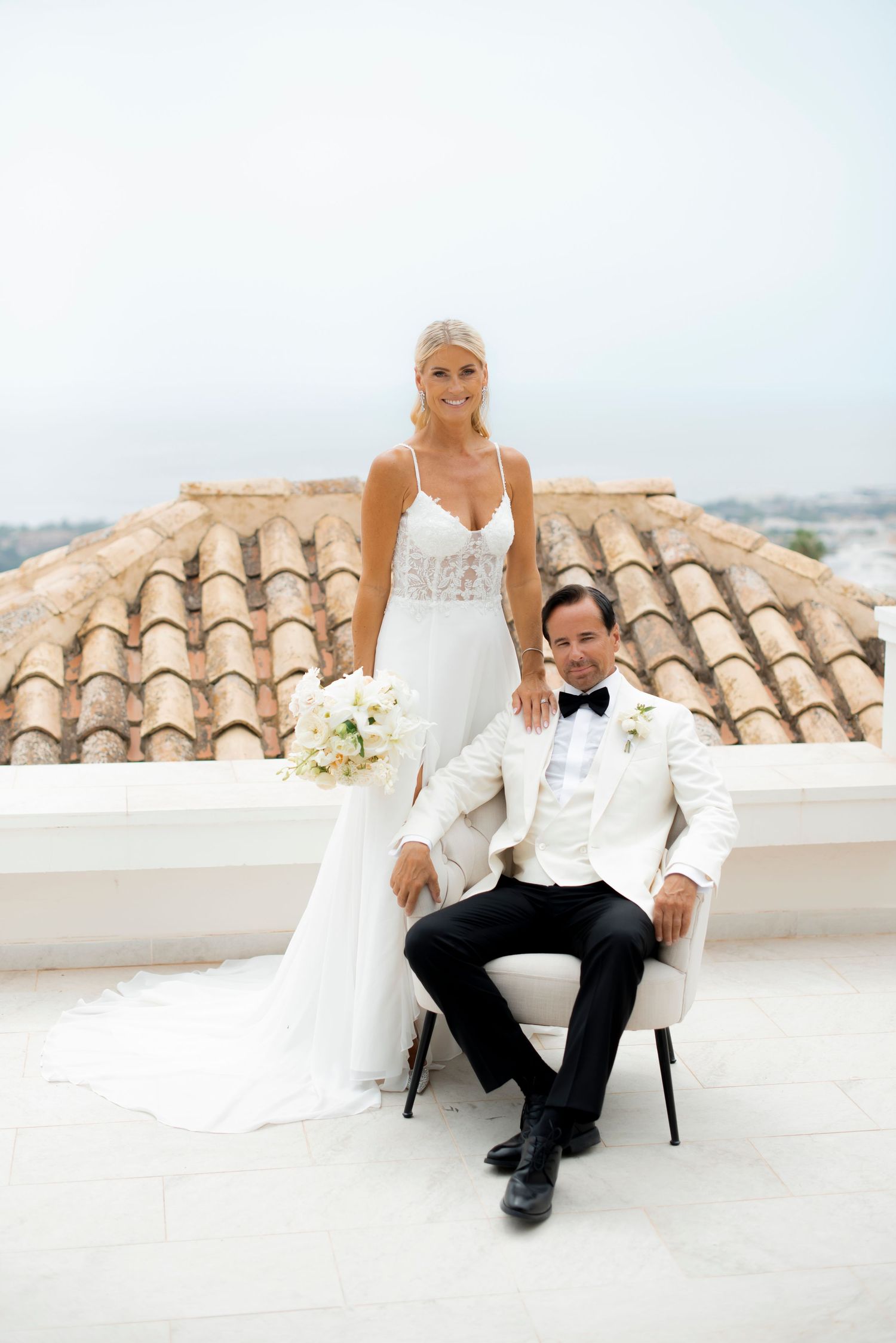 Bride in a lace gown holding a white bouquet stands beside a seated groom in a white tuxedo jacket and bow tie on a sunny terrace.