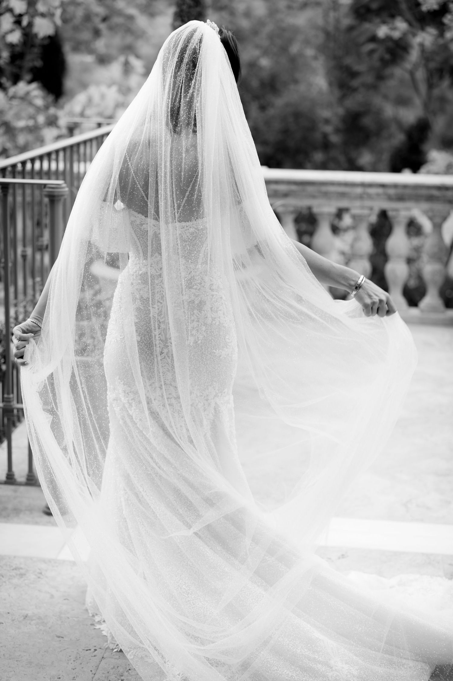 Black and white photo of a bride in a lace gown and long flowing tulle veil standing on a stone terrace with a balustrade.