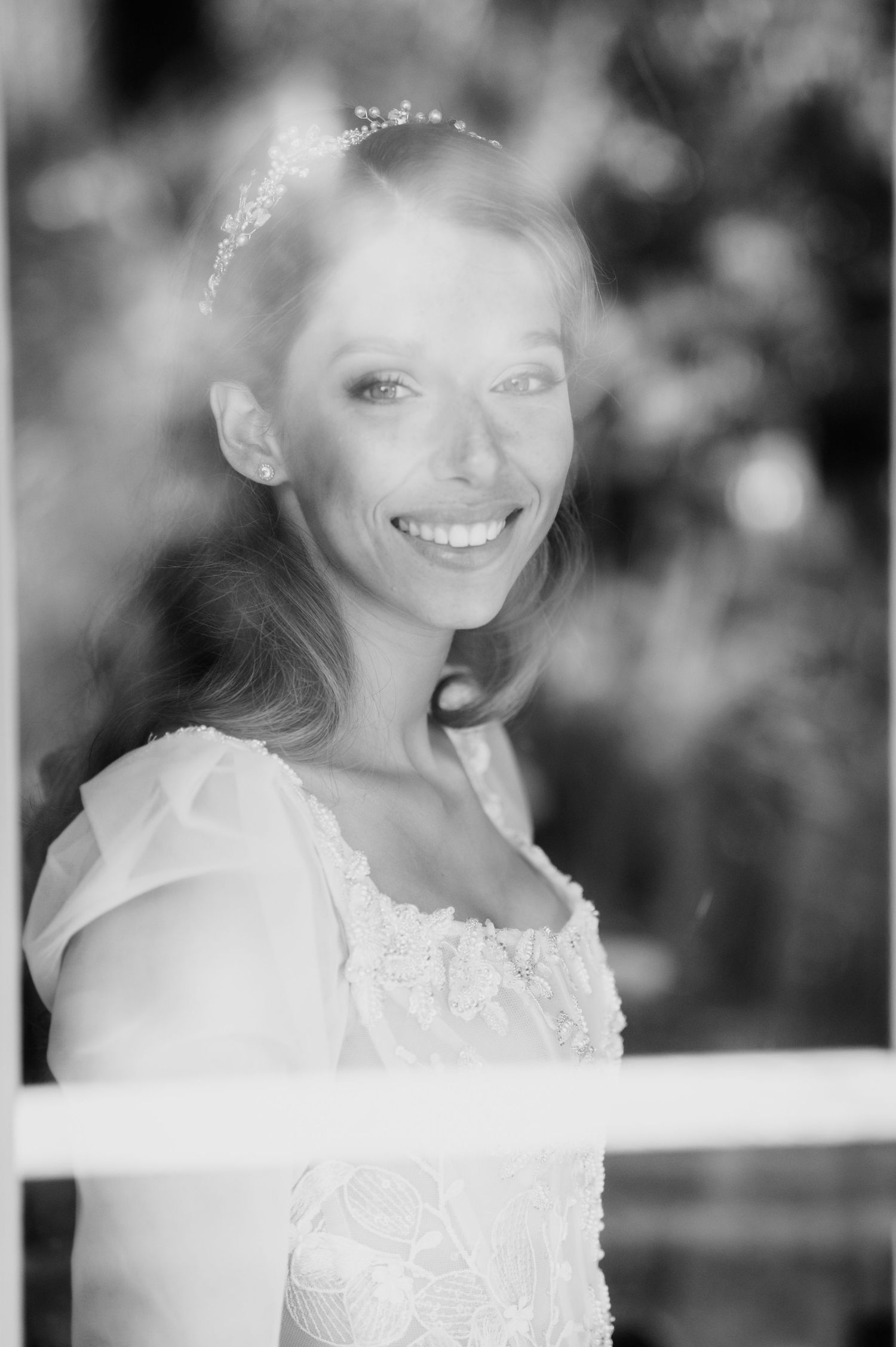 Black and white portrait of a smiling bride in a lace gown and beaded tiara, seen through a window with soft blurred greenery behind her.