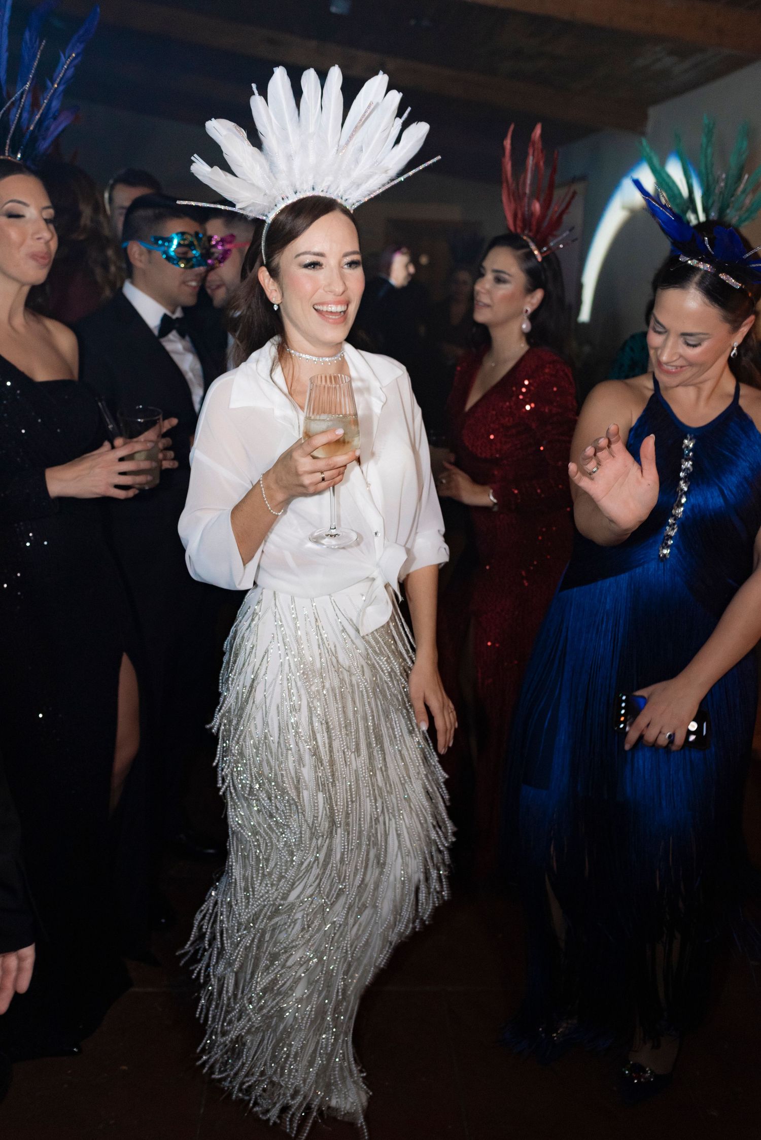 Smiling woman in a white feathered headdress and fringed skirt holds a champagne glass at a festive masquerade party with costumed guests.