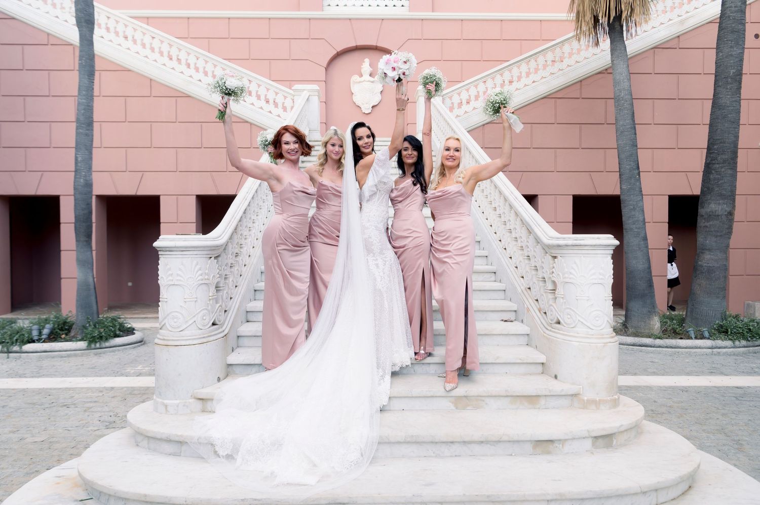 Bride in a white gown poses on a grand staircase with four bridesmaids in blush pink dresses holding bouquets against a pink building.