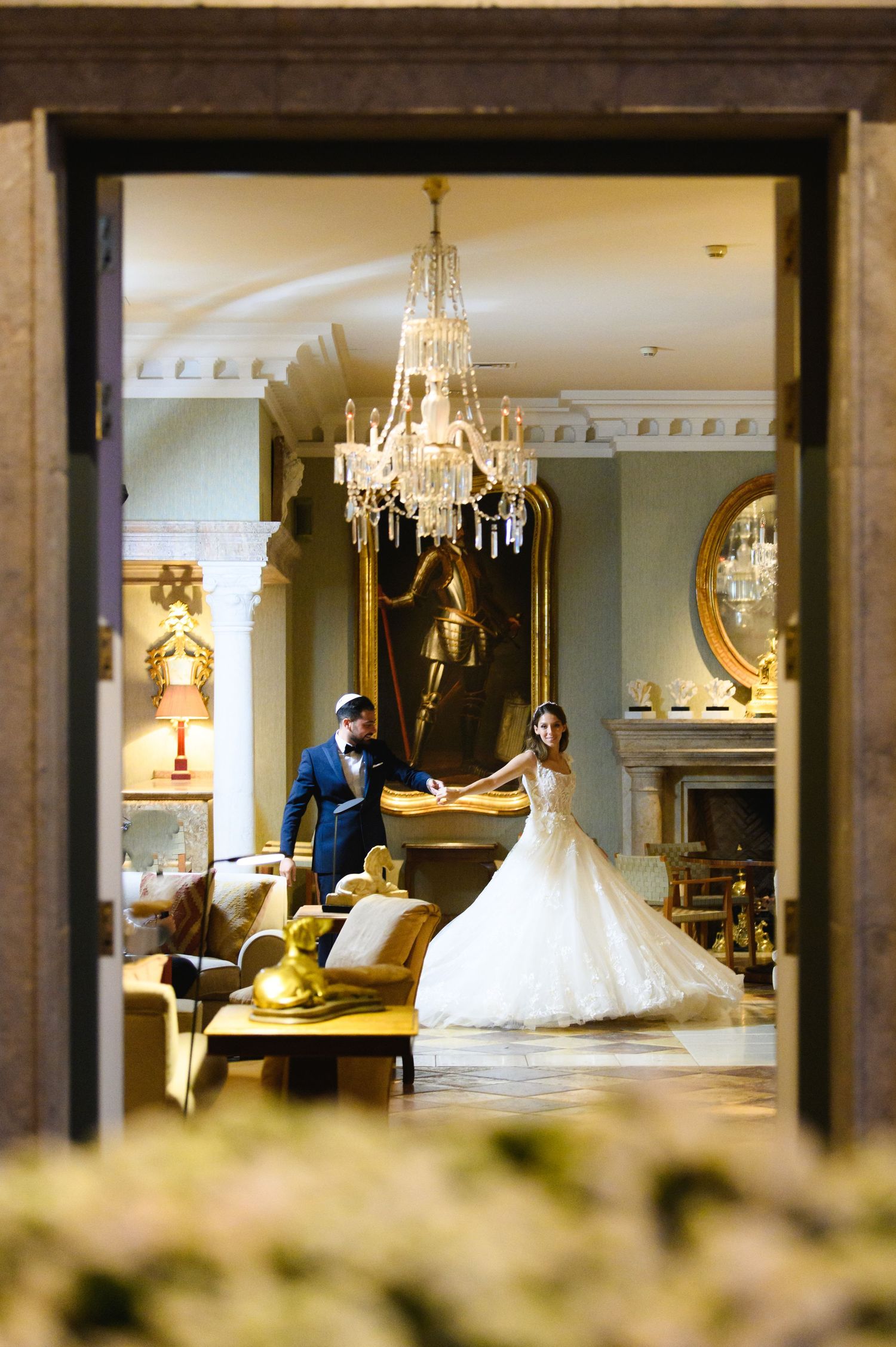 Bride in a ball gown and groom in a navy suit hold hands in an elegant hall with a crystal chandelier and ornate decor.