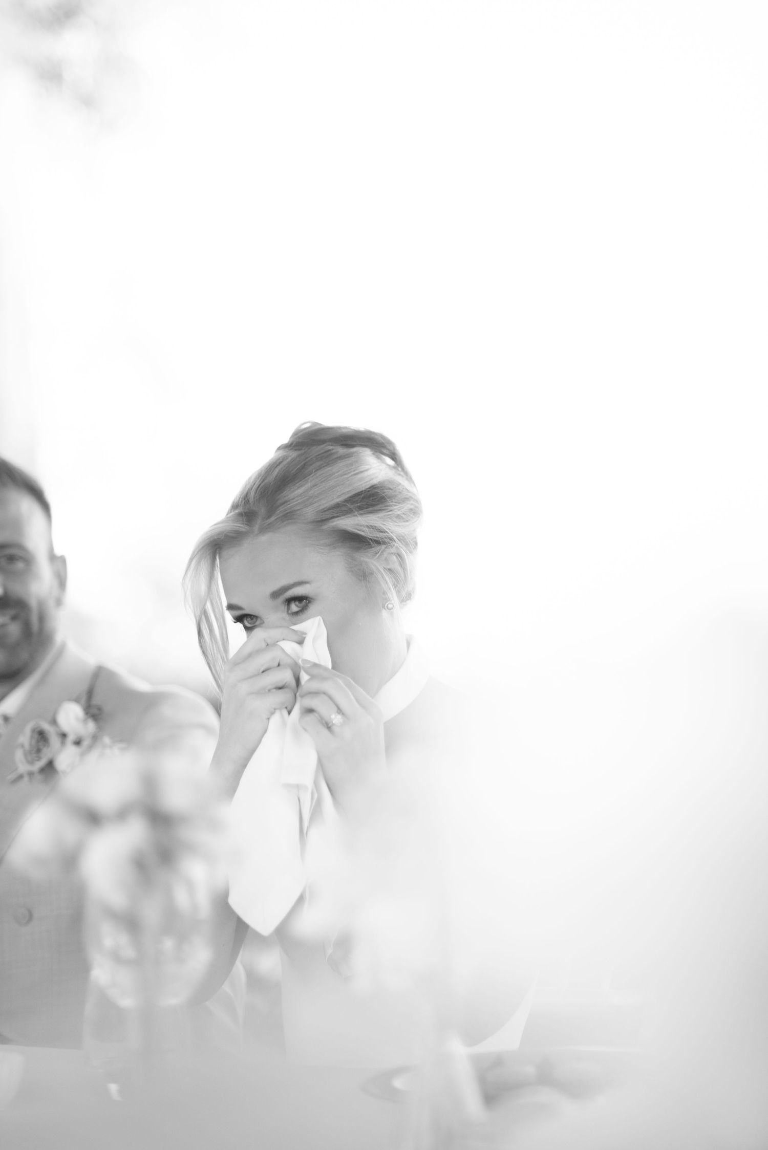 Black and white photo of a emotional bride dabbing her teary eyes with a napkin at a wedding reception, with a smiling groom beside her.