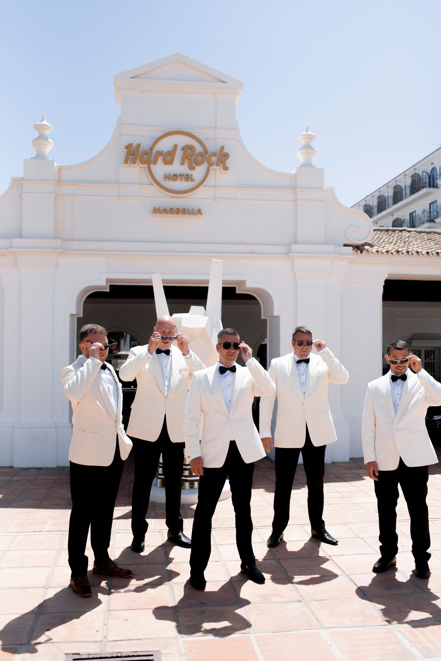 Five groomsmen in white tuxedo jackets and bow ties pose with sunglasses outside the Hard Rock Hotel Marbella on a sunny wedding day.