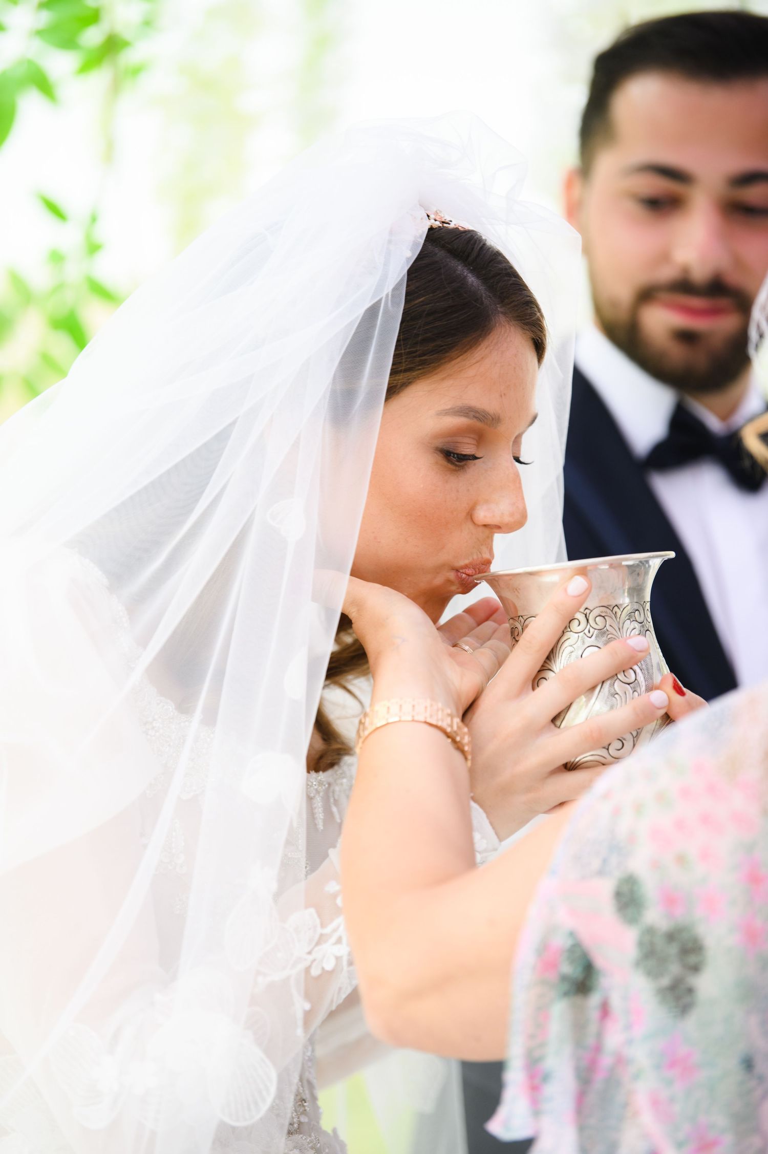 Bride in a flowing white veil sips from an ornate silver goblet during a wedding ceremony while the groom looks on.