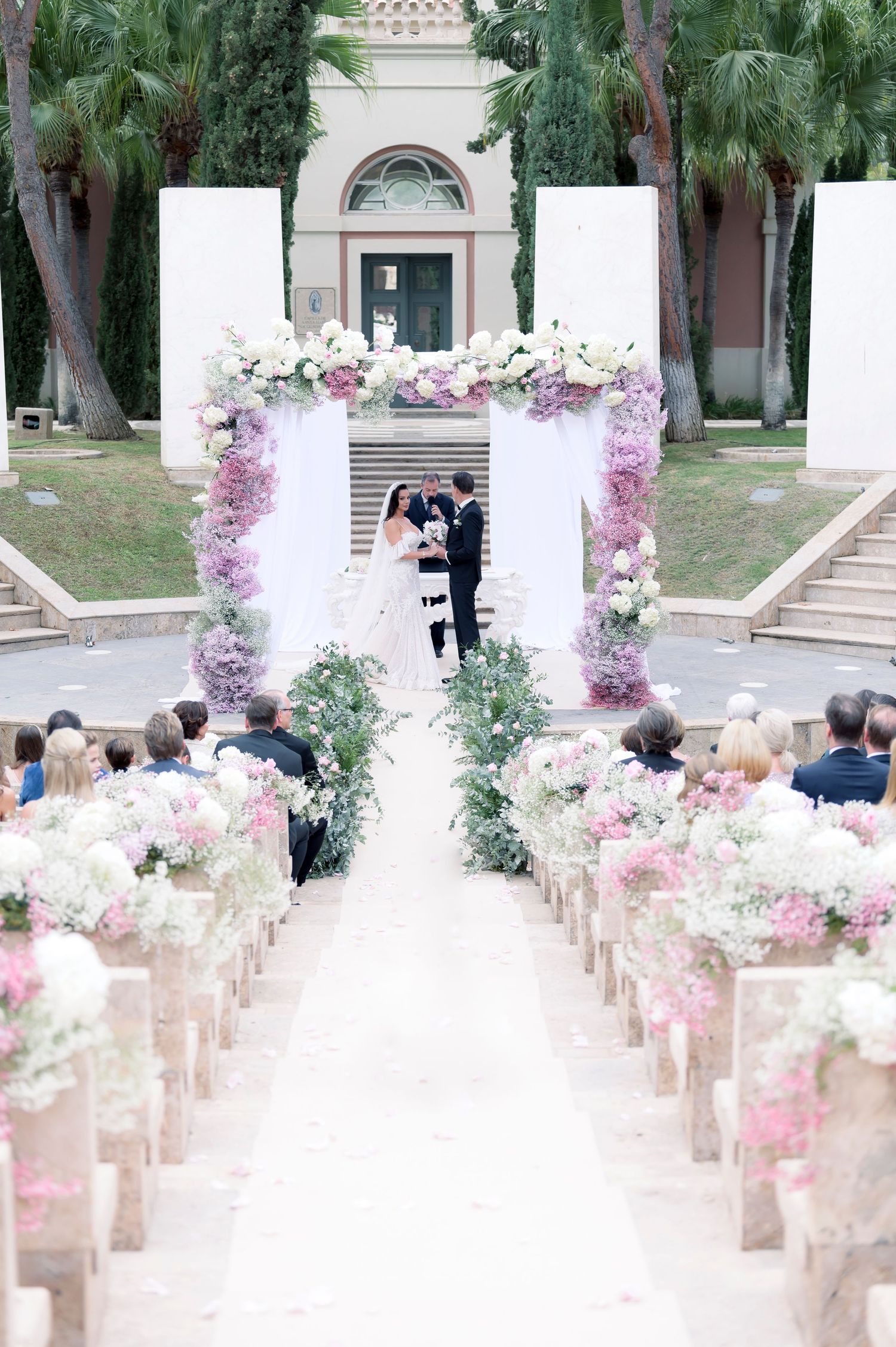 Bride and groom exchange vows beneath a floral arch at an outdoor wedding ceremony, with guests seated along an aisle lined with flowers.