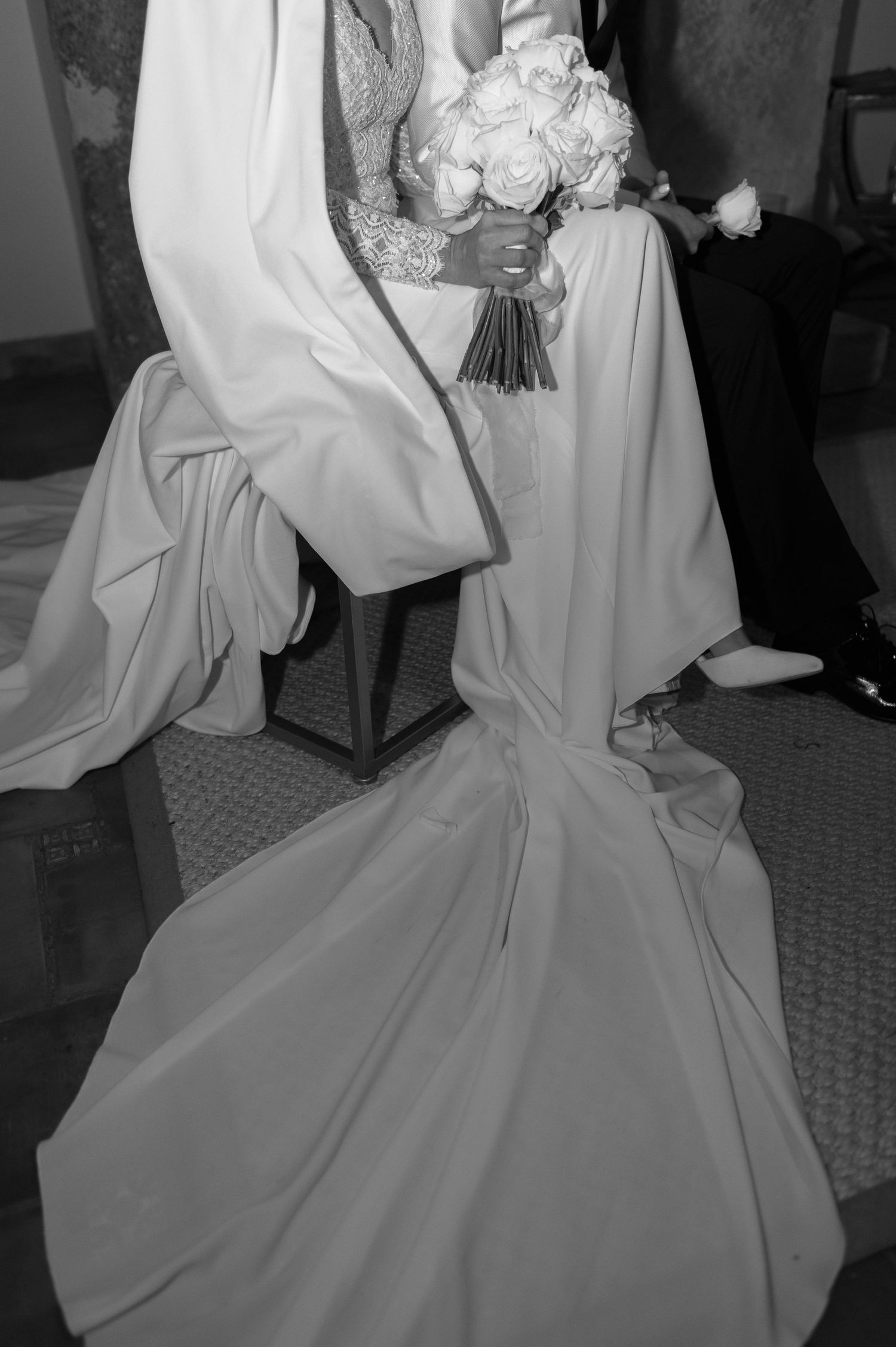 Black and white photo of a seated bride in a flowing satin gown with a long train, holding a bouquet of white roses.