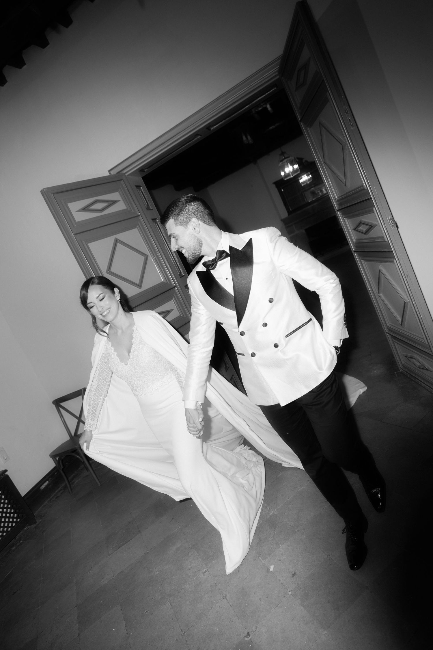 Black and white photo of a smiling bride in a flowing cape gown holding hands with a groom in a white tuxedo jacket and bow tie.