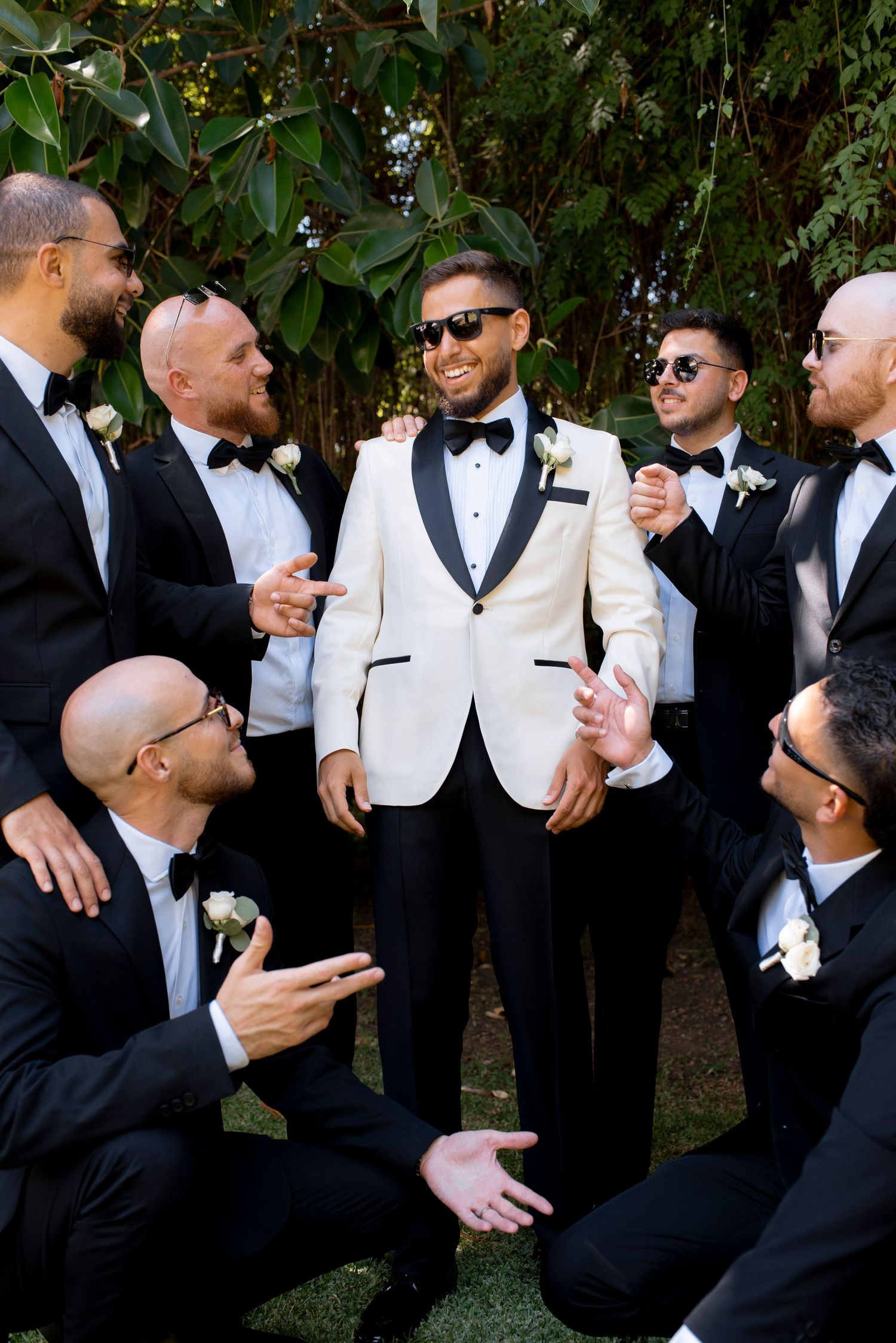 Groom in a white dinner jacket laughs with groomsmen in black tuxedos and sunglasses, posing playfully outdoors among green trees.