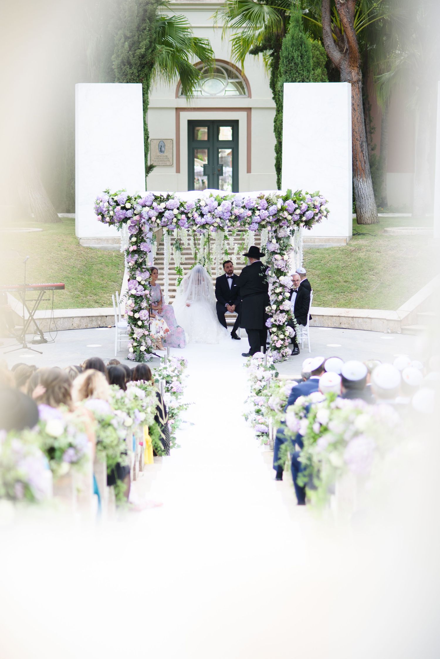 Outdoor wedding ceremony under a purple floral chuppah with guests seated along a flower-lined aisle.