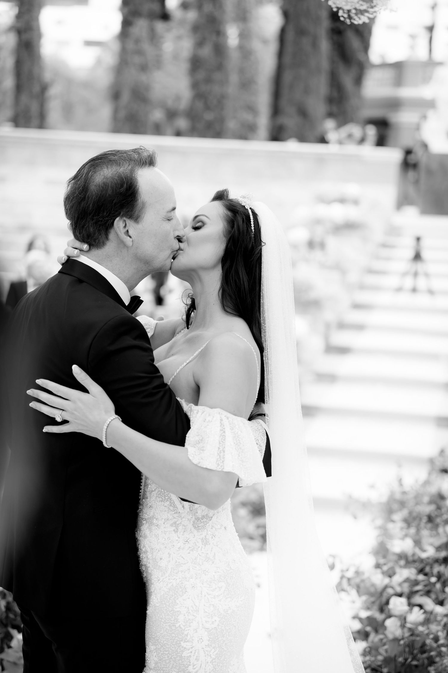 Black and white photo of a bride in a lace gown and veil kissing her groom in a tuxedo at an outdoor wedding ceremony.