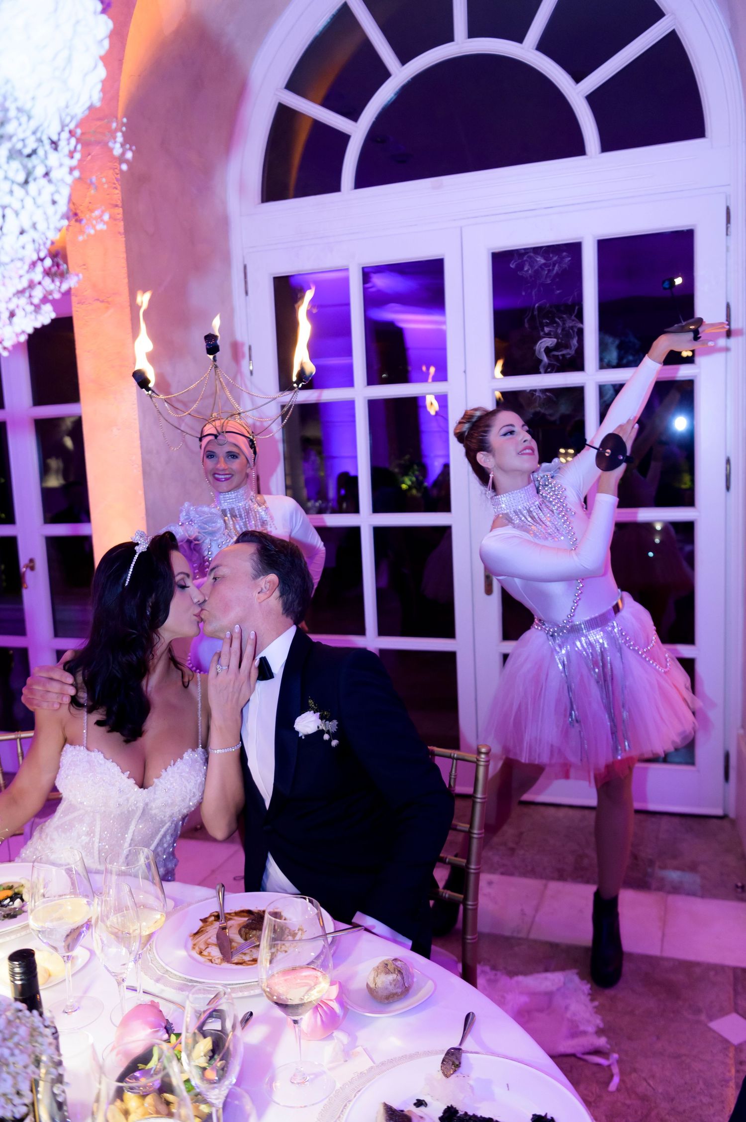 Bride and groom share a kiss at their reception table while costumed performers with flaming headpieces entertain nearby.