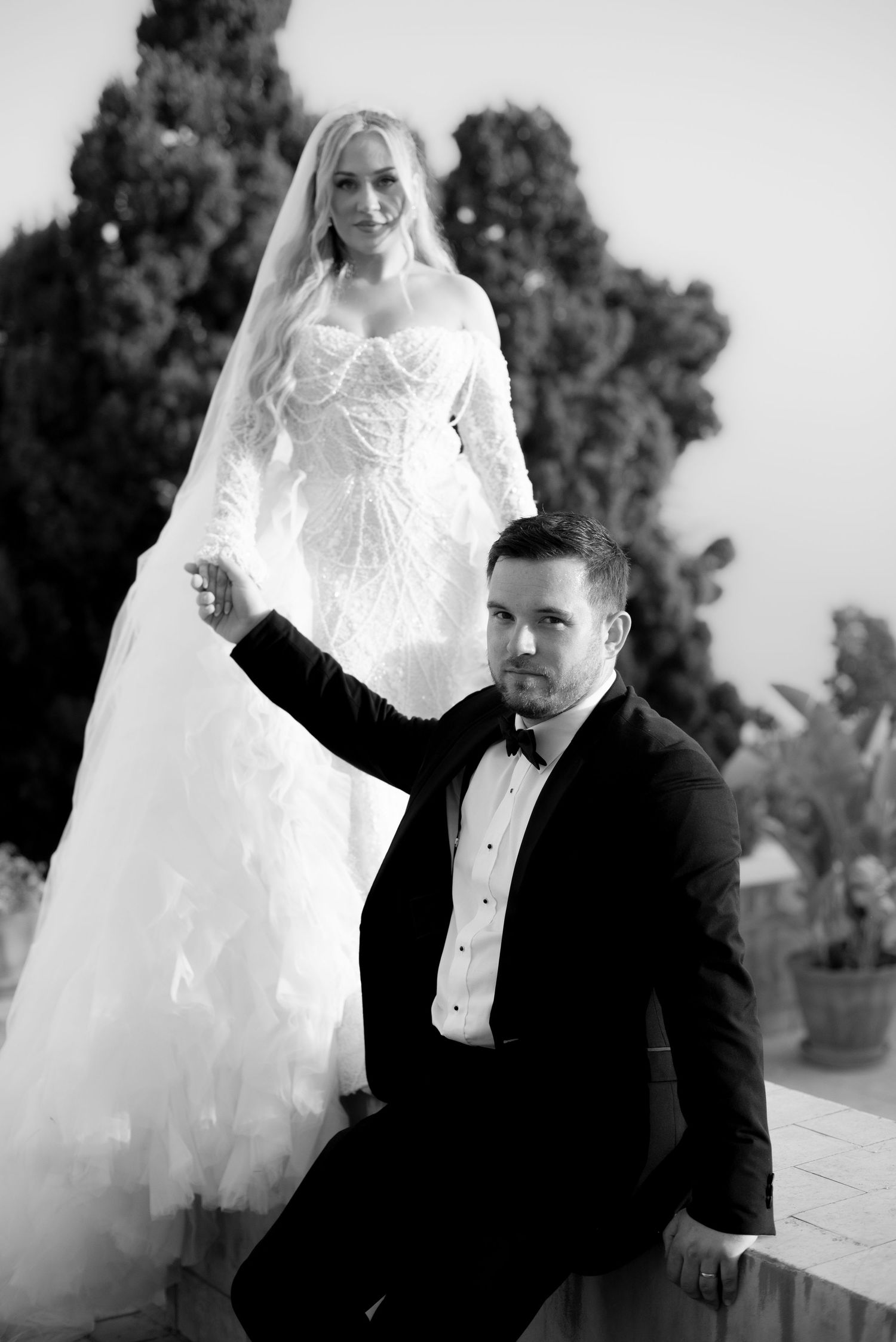 Black and white wedding portrait of a bride in a lace gown with long veil standing behind a groom in a tuxedo and bow tie.