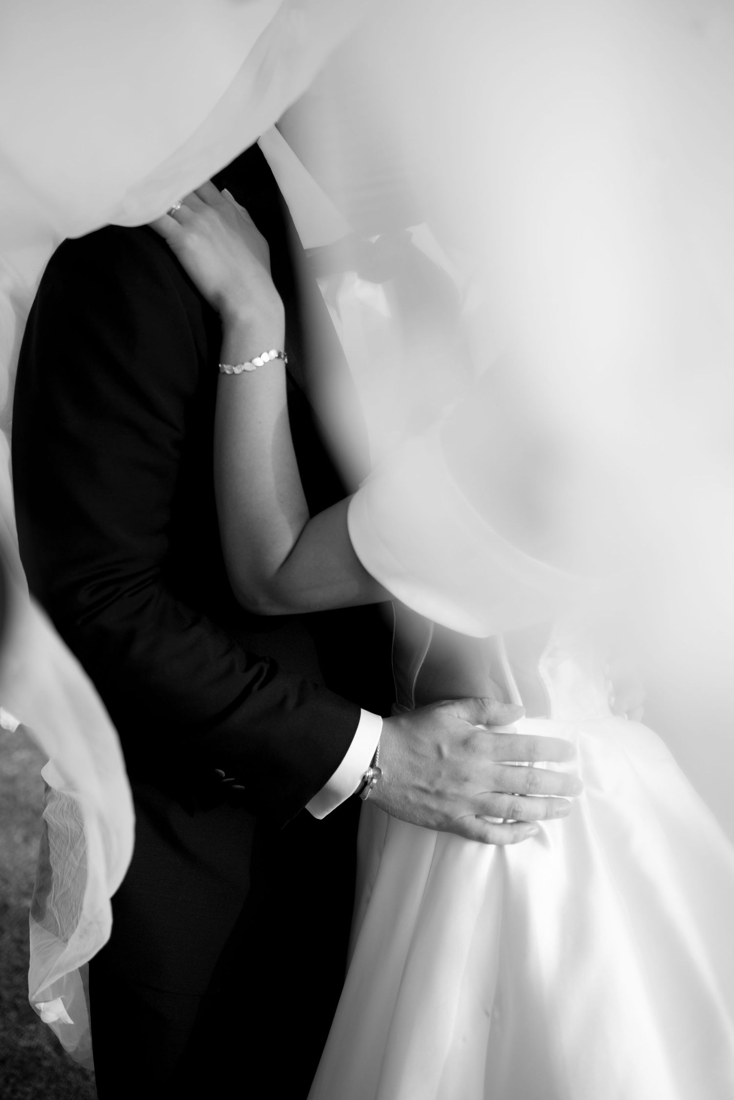 Black and white photo of a bride and groom embracing under a sheer veil, her bracelet and rings visible as her hand rests on his shoulder.