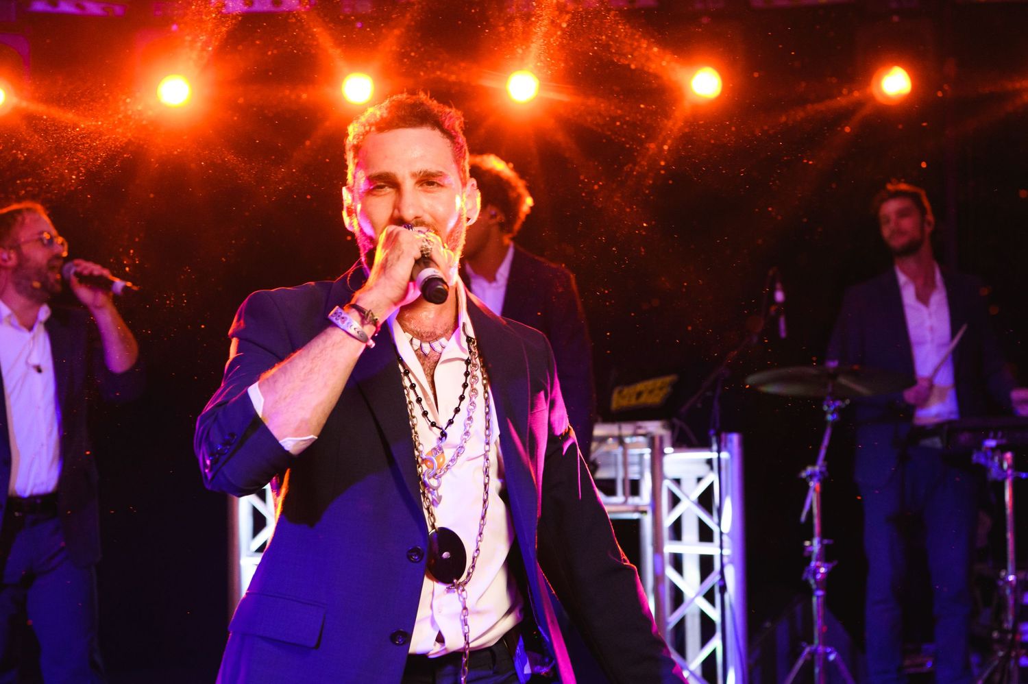 Male singer in a navy blazer and layered necklaces performs into a microphone on stage with a live band behind him.