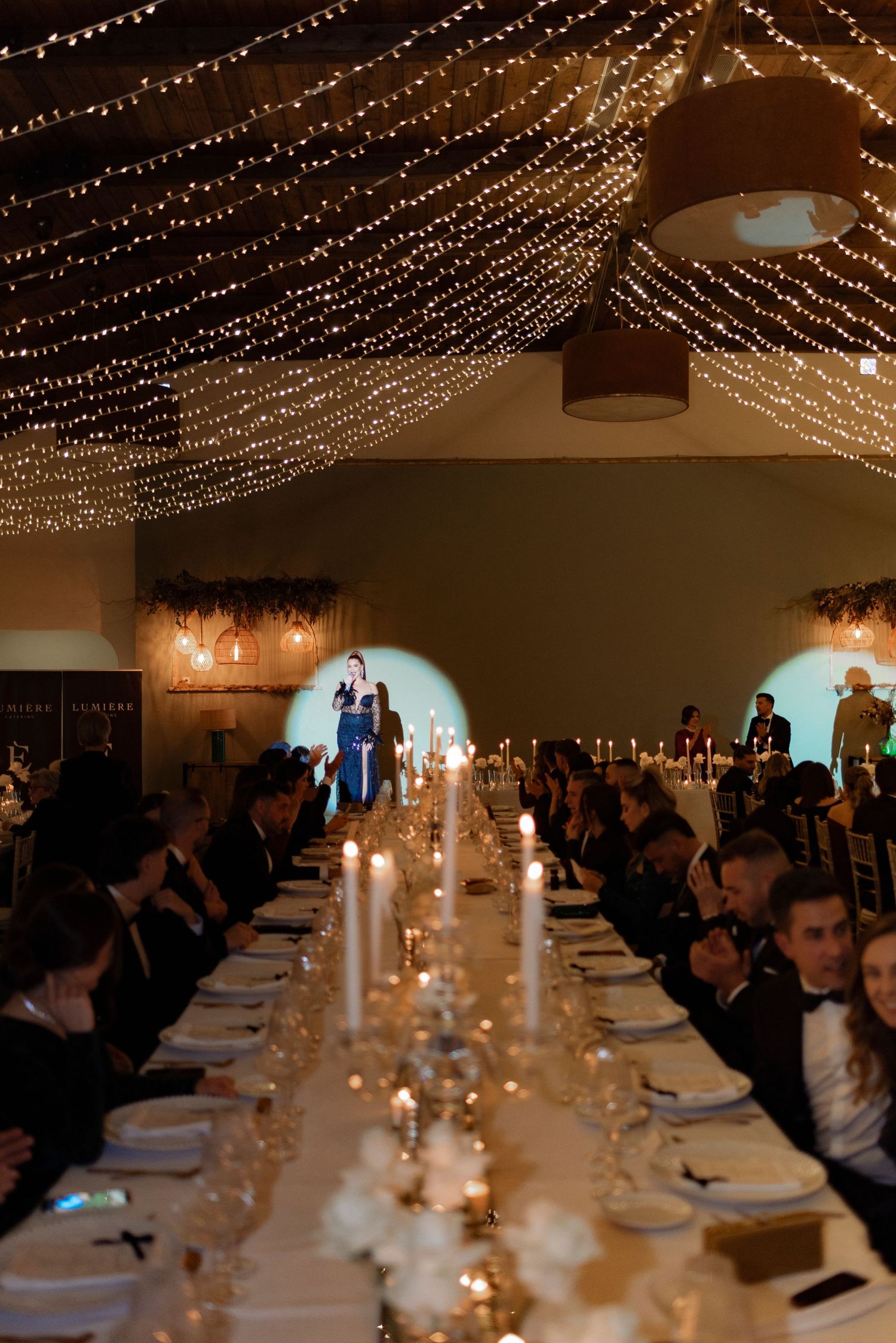 Wedding guests in formal attire dine at a long candlelit table beneath canopy string lights while a woman in blue speaks in a spotlight.