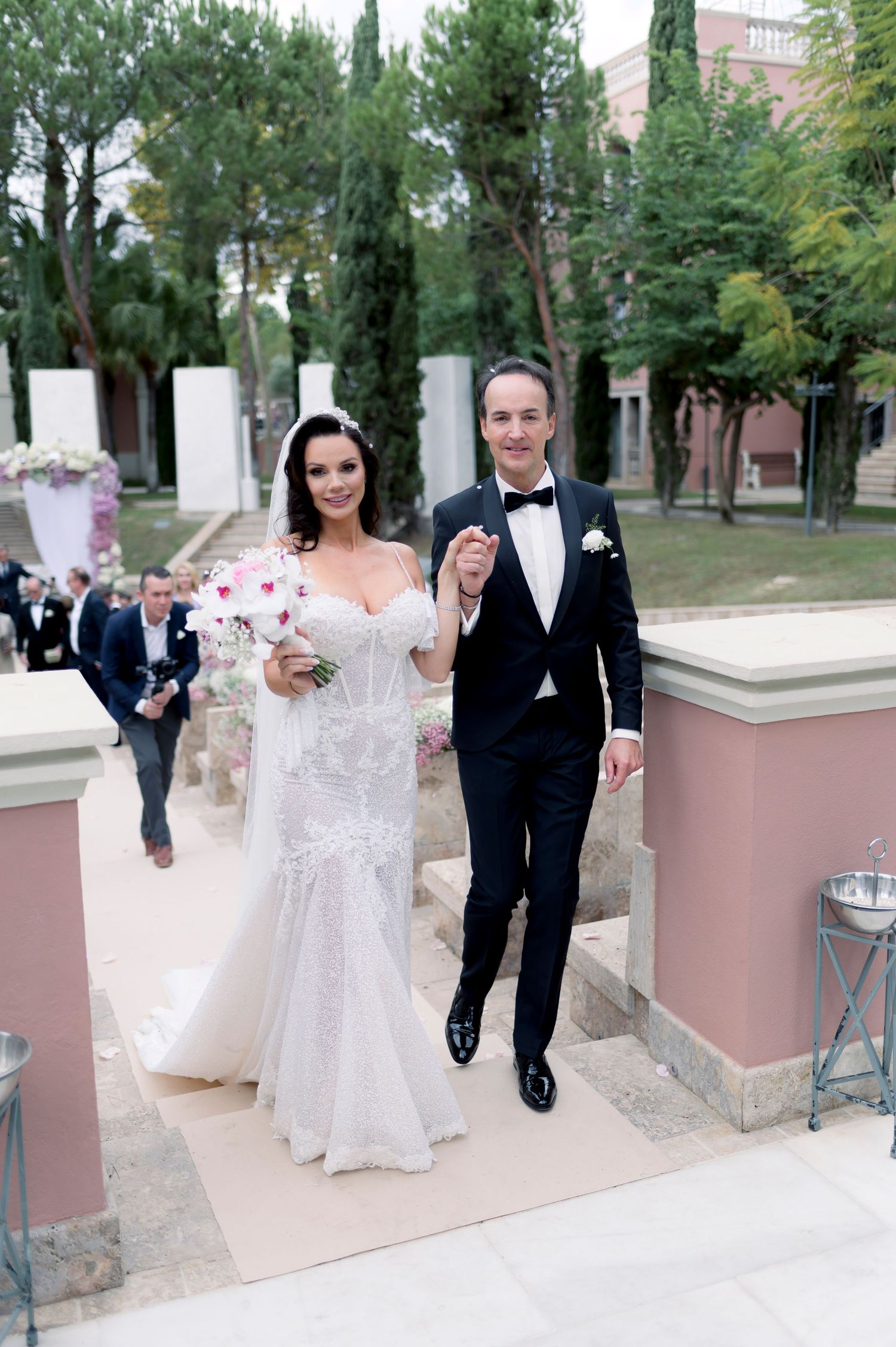 Bride in a lace wedding dress holding a floral bouquet walks hand in hand with a groom in a black tuxedo outdoors.