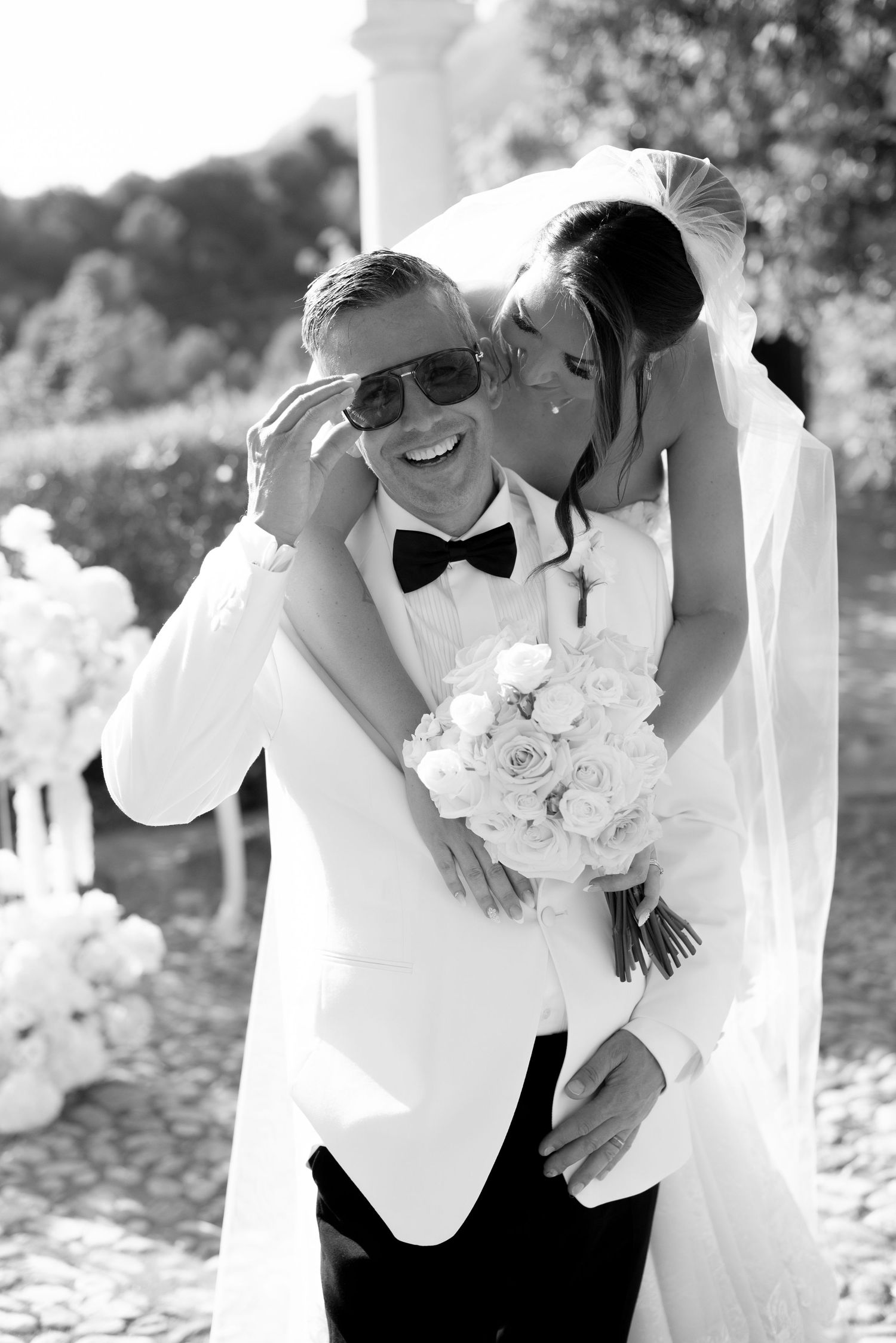 Black and white wedding photo of a smiling groom in sunglasses and bow tie holding a rose bouquet as the bride in a veil kisses his cheek.