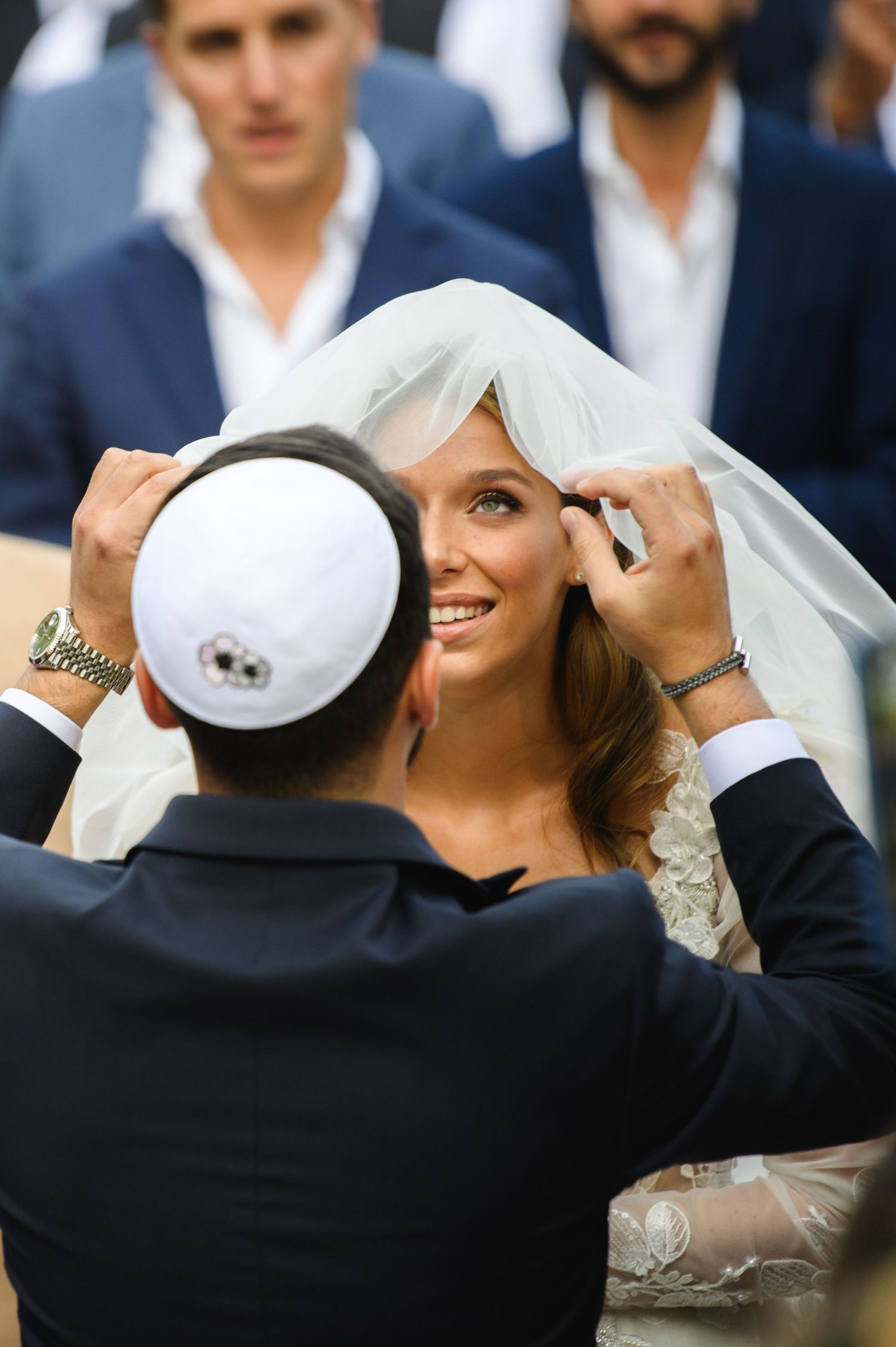 A groom in a white kippah lifts the veil of a smiling bride during a Jewish wedding ceremony as guests in suits look on.